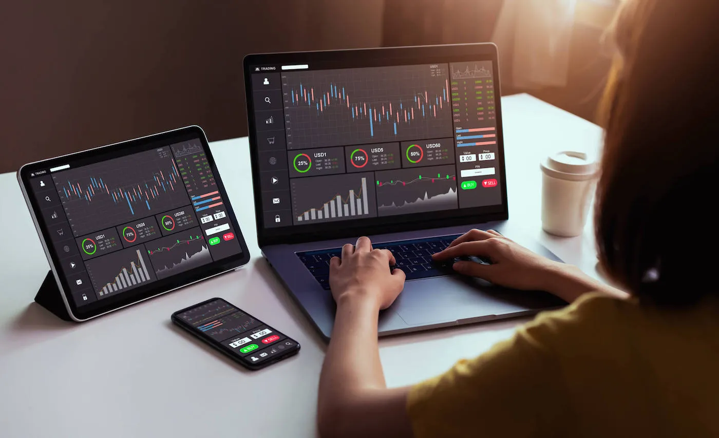 A woman types on a laptop that is next to a tablet and phone all showing the same stock chart.