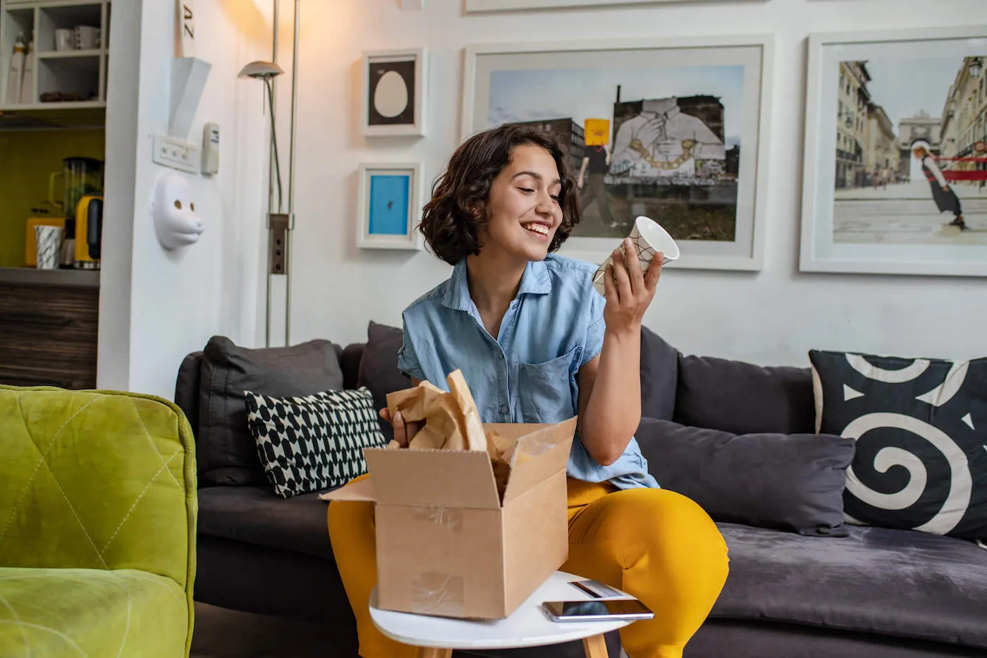 A young women wearing yellow pants is sitting on the couch and smiling while she opens a package containing a white cup.