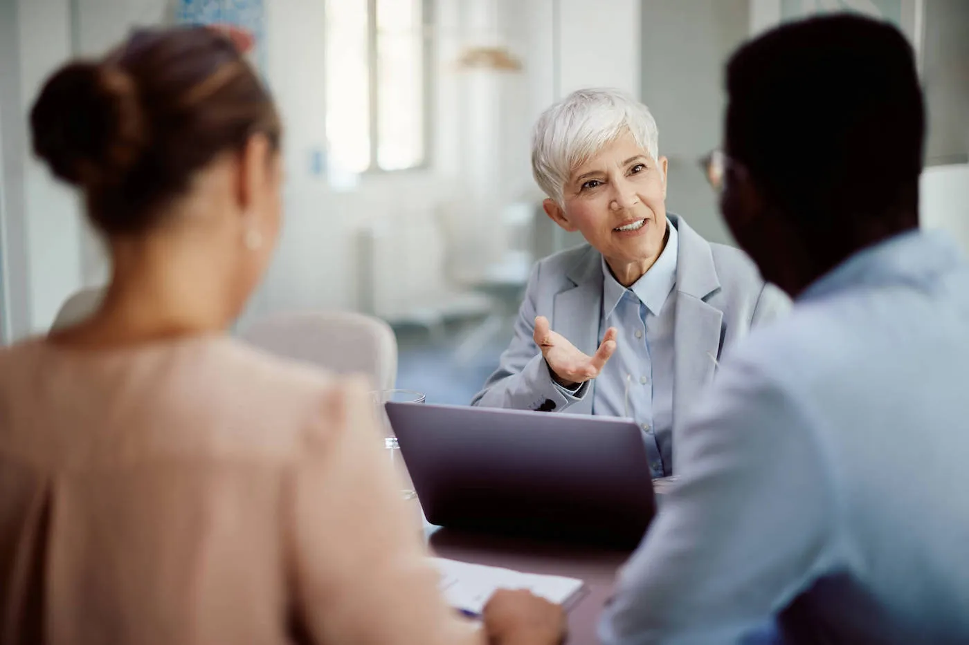 A women wearing a gray suit is talking with a couple across the table while she has her laptop open.