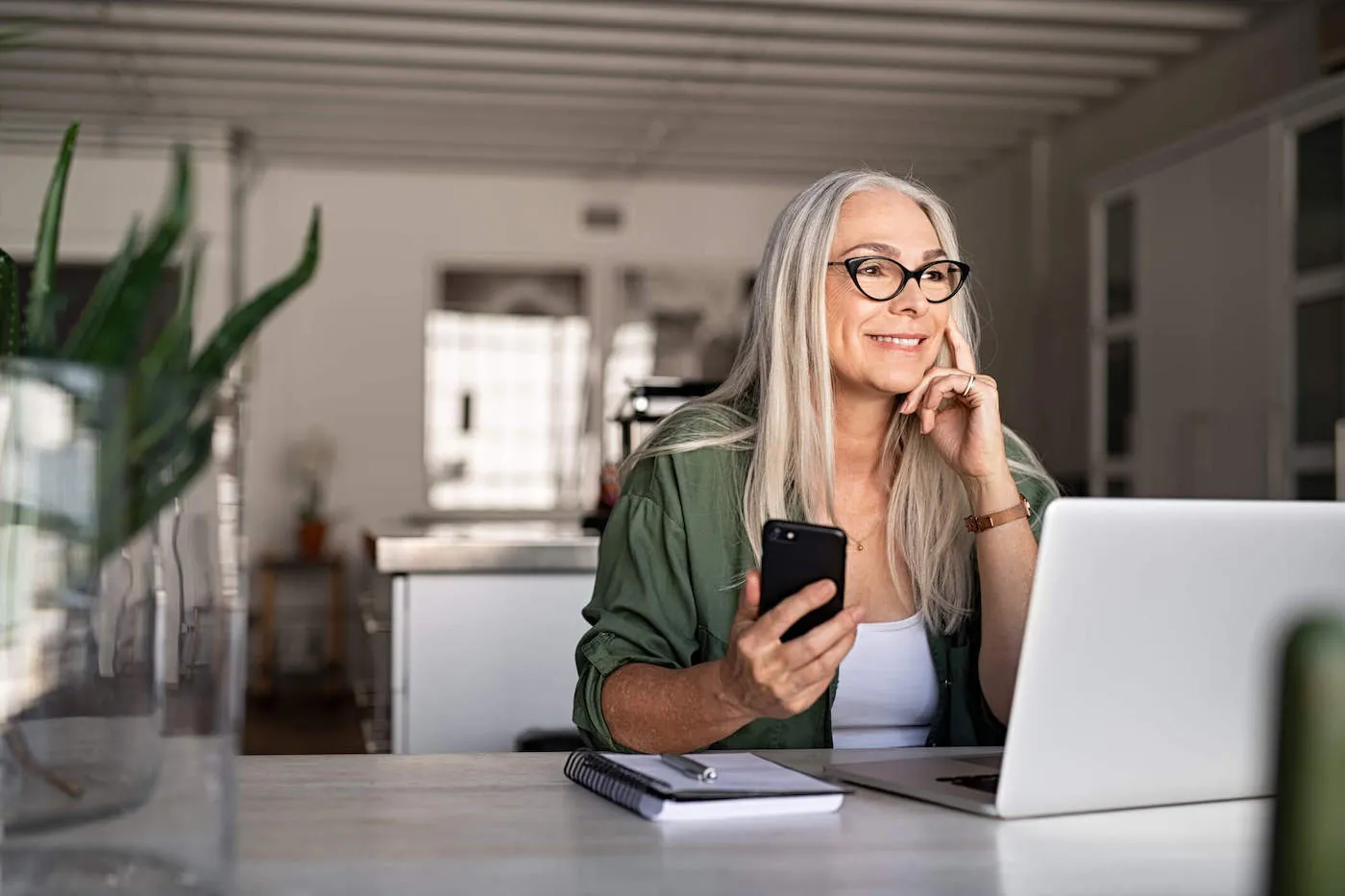 A women wearing a green jacket and black glasses smiles as she looks out her kitchen window while holding her phone.