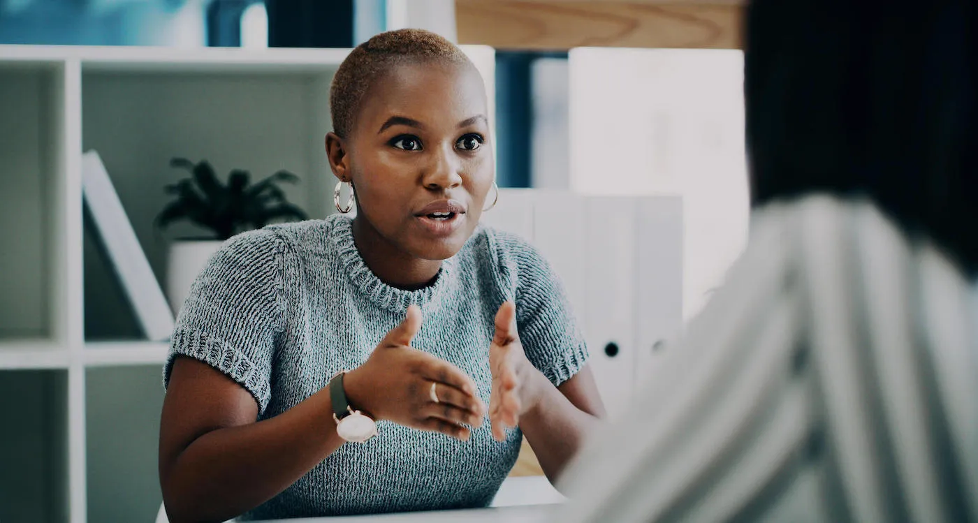 A women wearing a gray sweater and hoop earrings talks to a person across the table at the office.