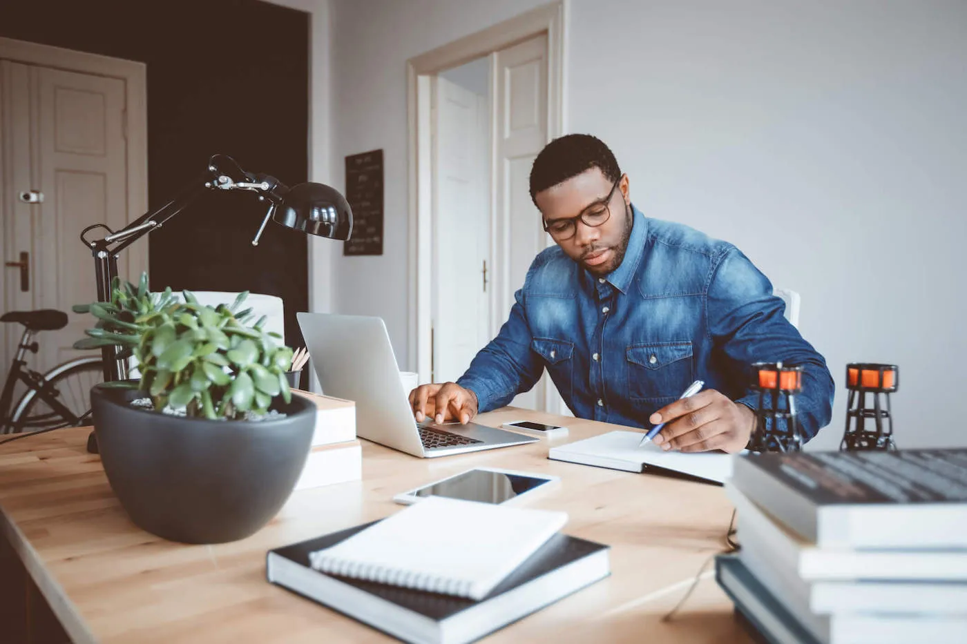 A young man wearing a blue shirt writes on a journal while also using his laptop computer at the desk.