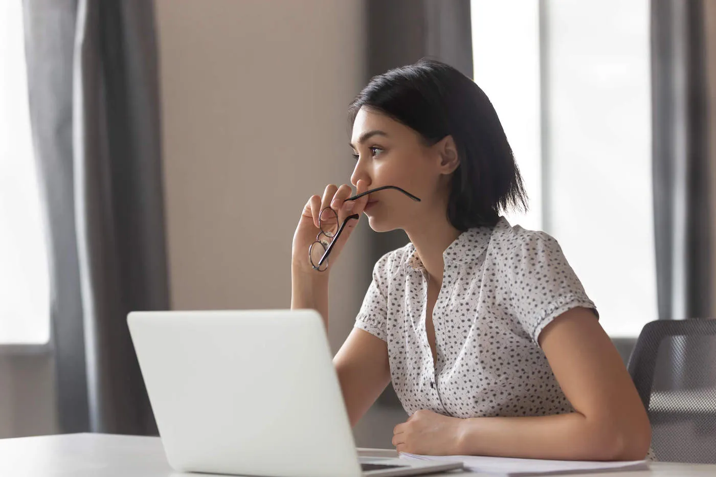 A young woman holds her glasses while she looks away from her computer.