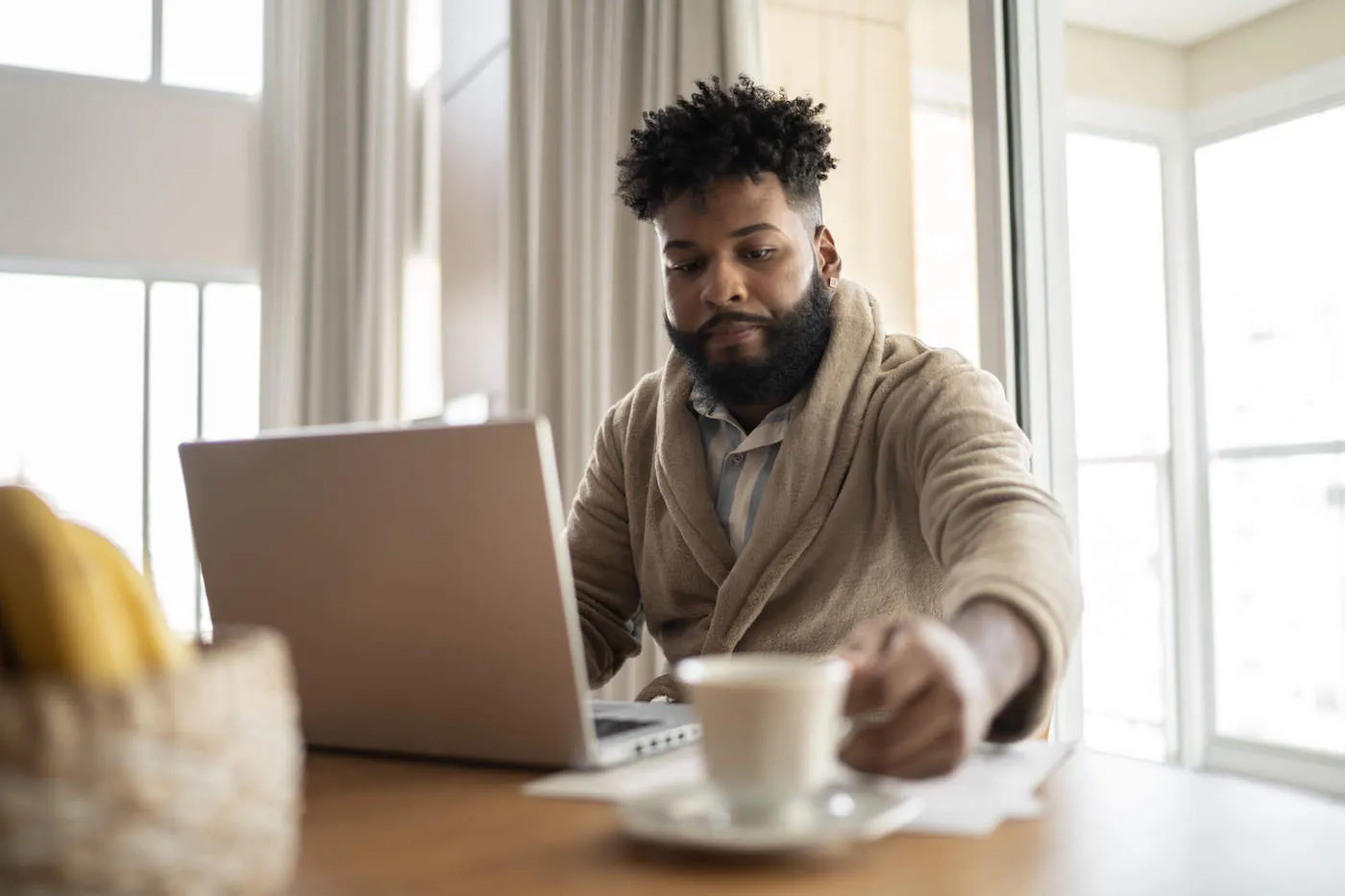 African American man in bathrobe reaching for cup while seated in front of open laptop.