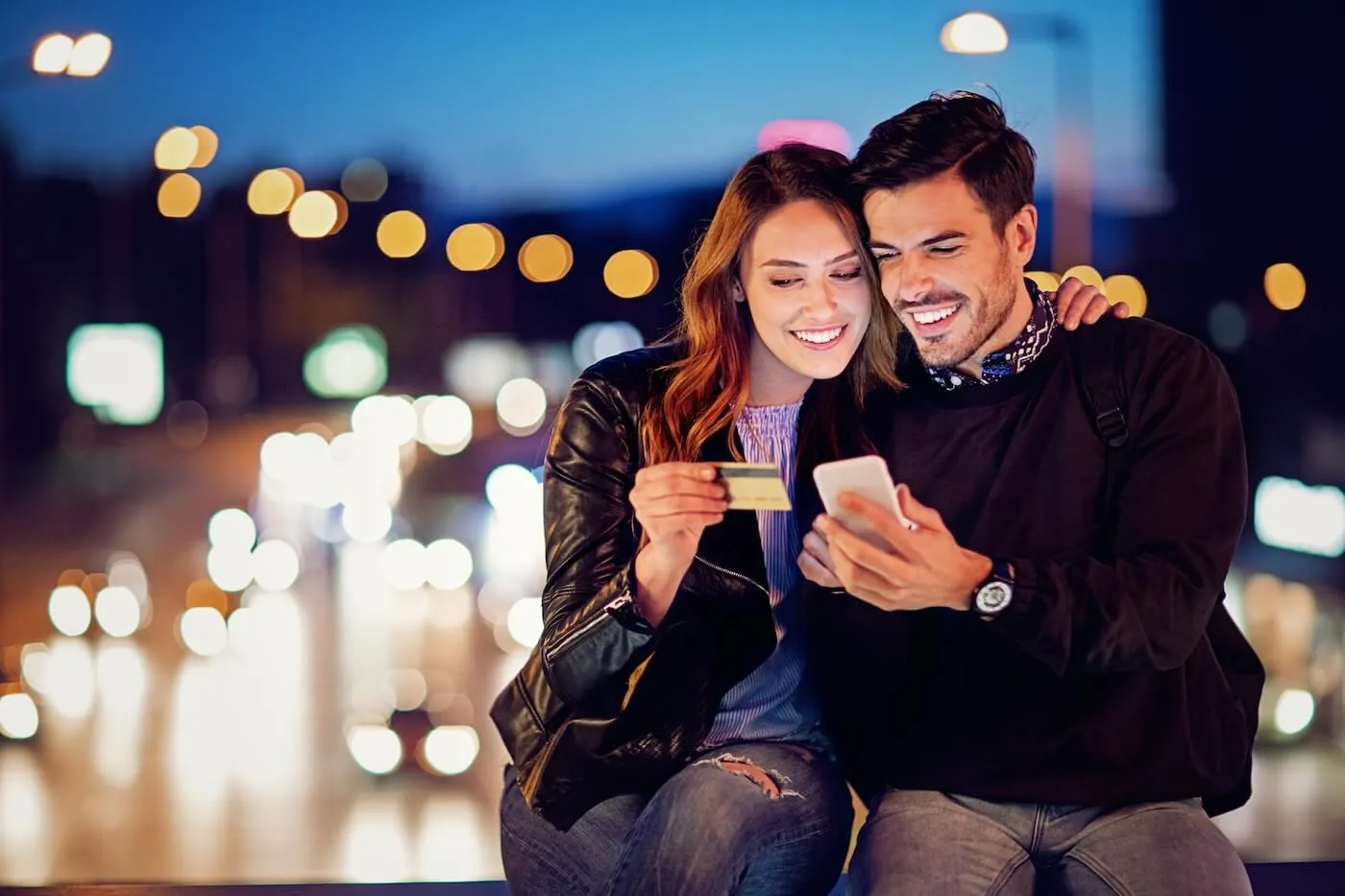 Happy young couple sitting closely together outdoors at night, with the woman holding a payment card and the man holding a smartphone, against a blurred city street background with glowing streetlights and traffic lights.