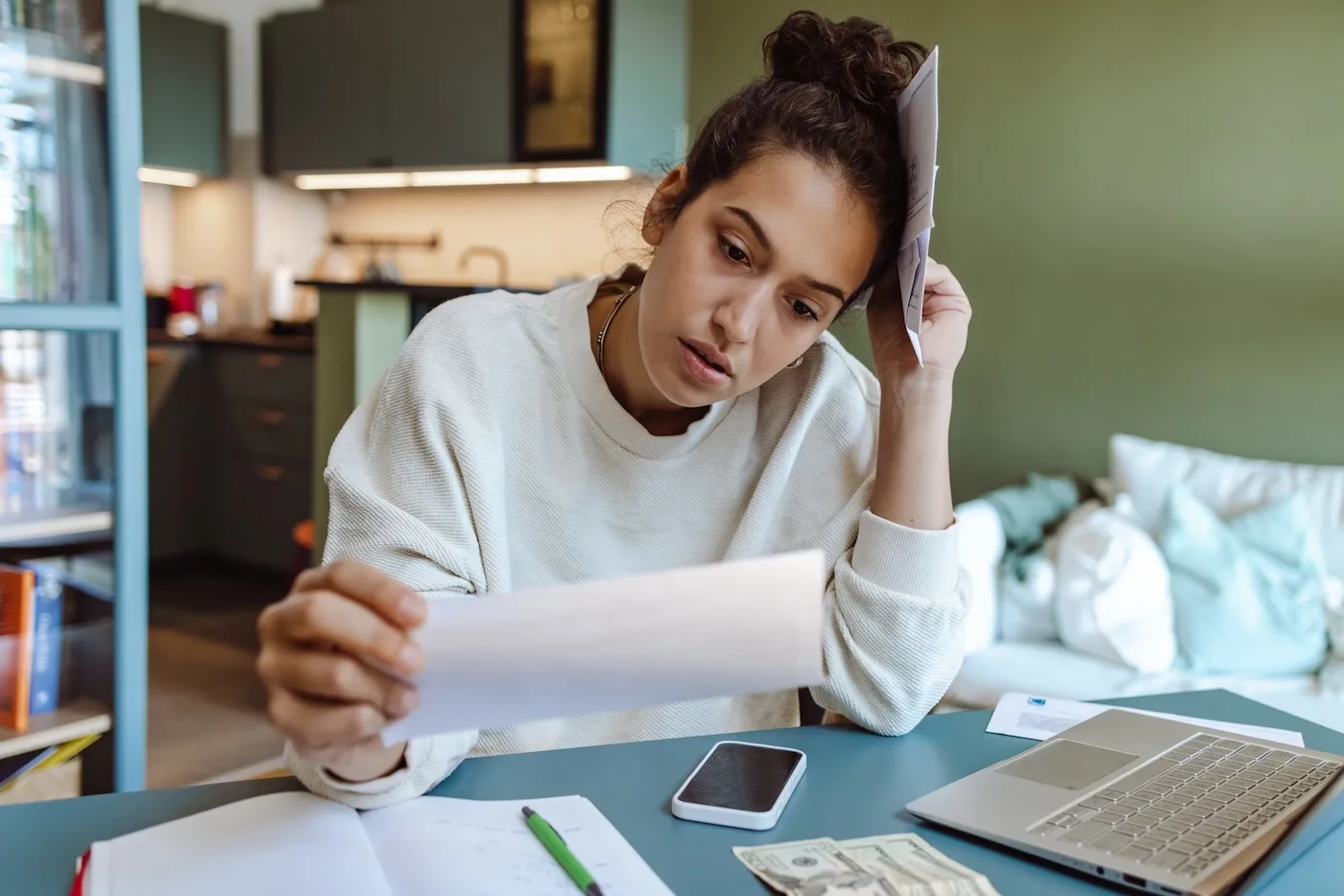 Young woman in a beige sweater sitting at a table and going through paperwork. She is holding unpaid bills.