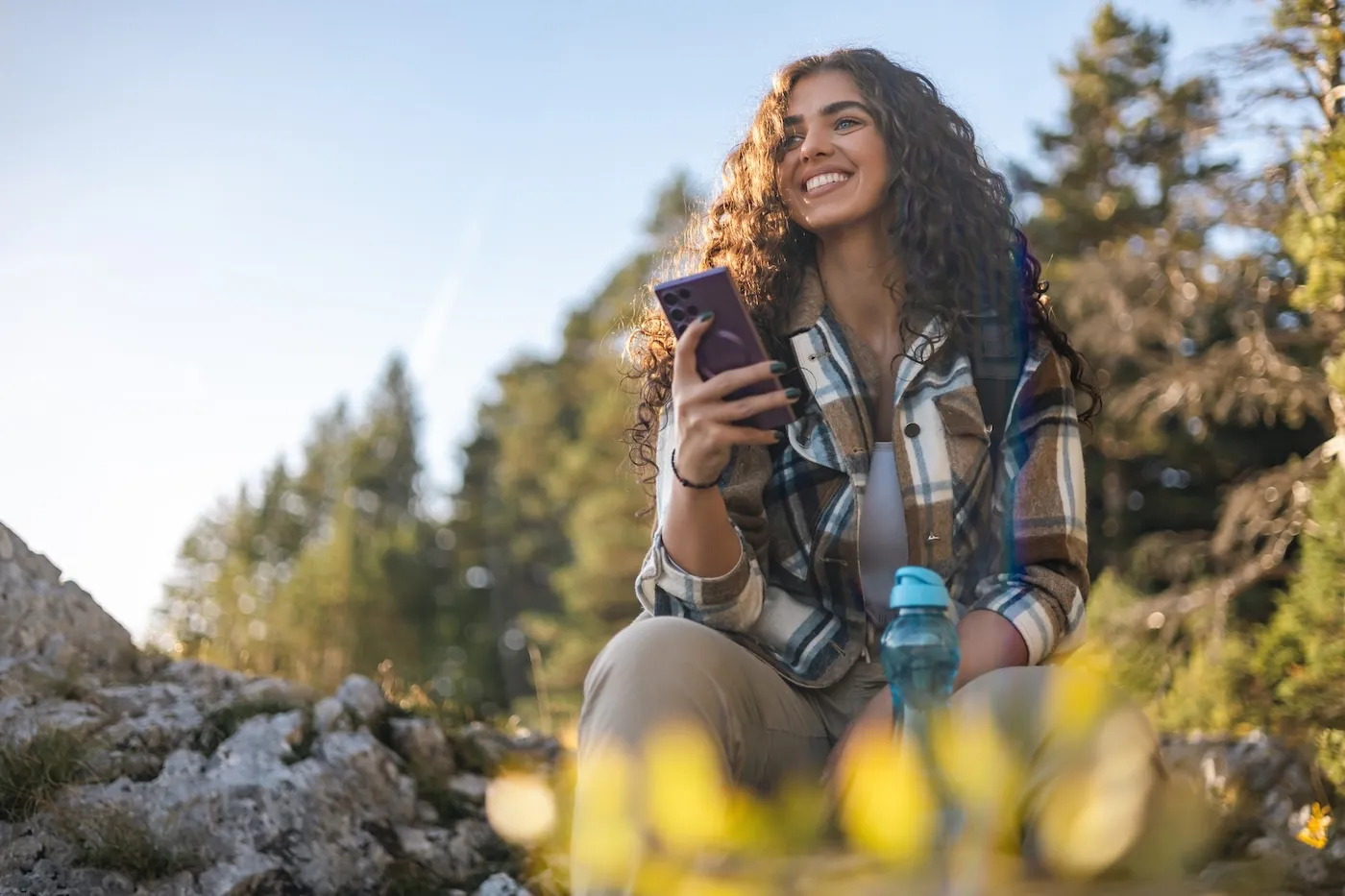 A smiling woman with curly hair, dressed in a plaid shirt, using her phone while sitting comfortably on rocks amidst a scenic forest setting.