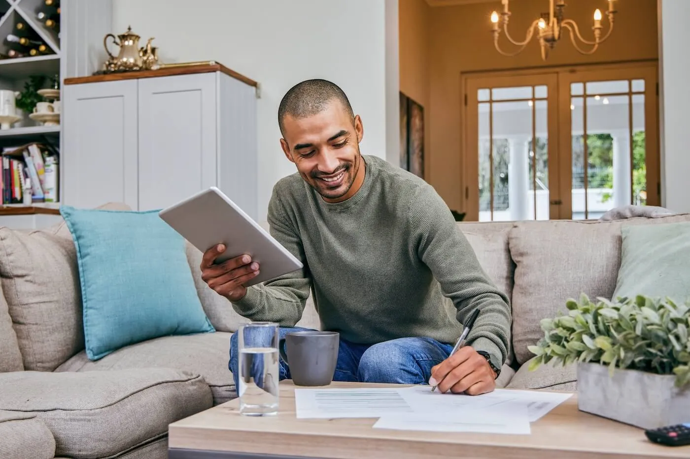 A smiling man sitting on the couch and checking an investing app on a tablet while making notes