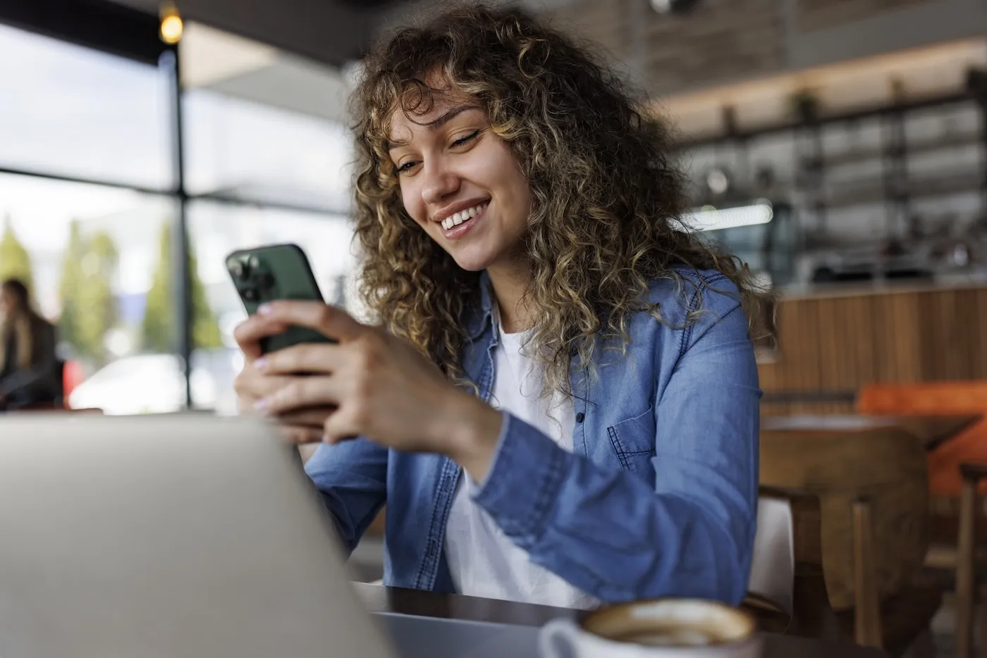 Young smiling woman using mobile phone while working on a laptop at a cafe.