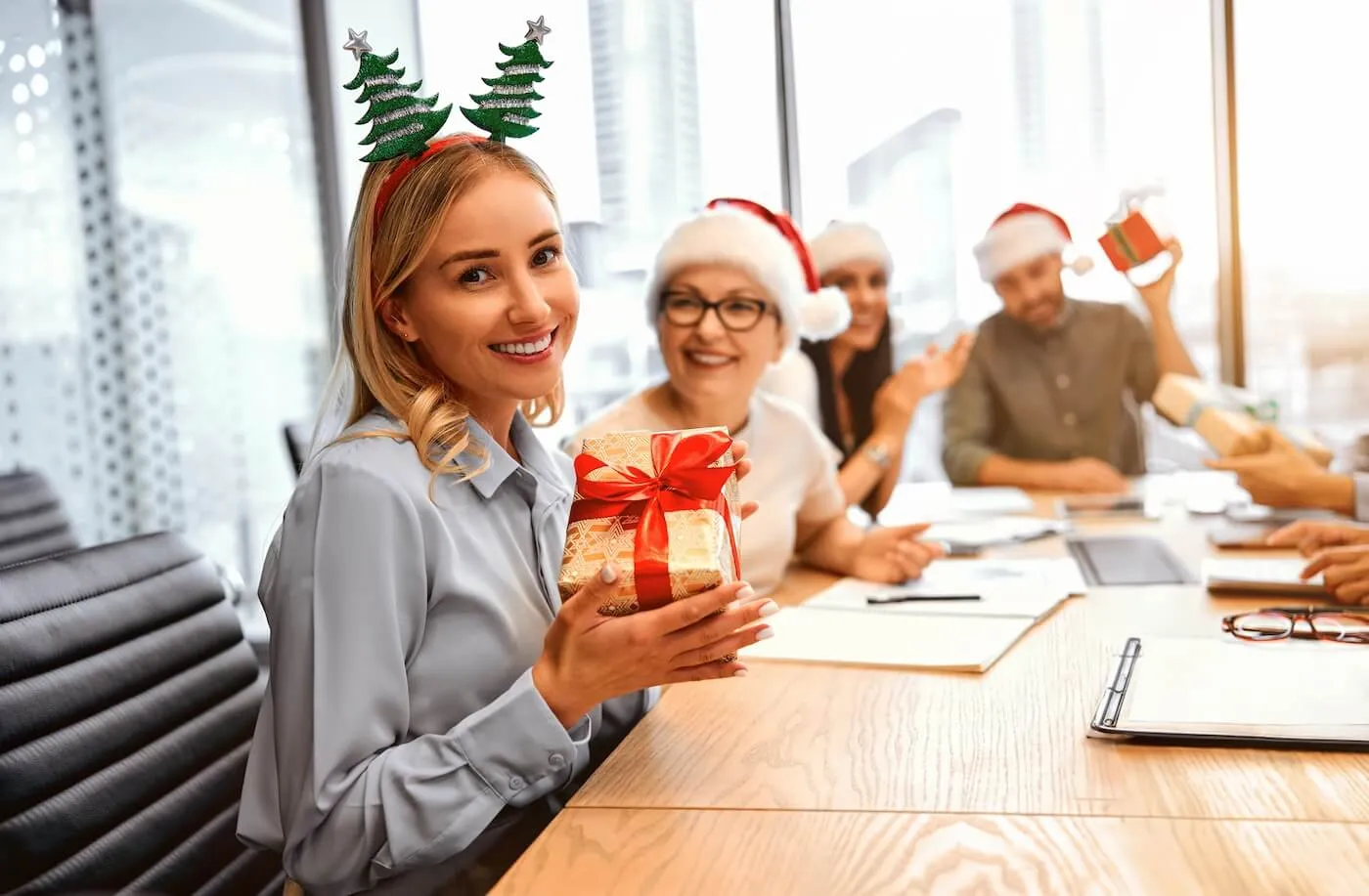 A young woman wearing a Christmas hairband is holding a present box in the office, surrounded by by coworkers in festive outfits