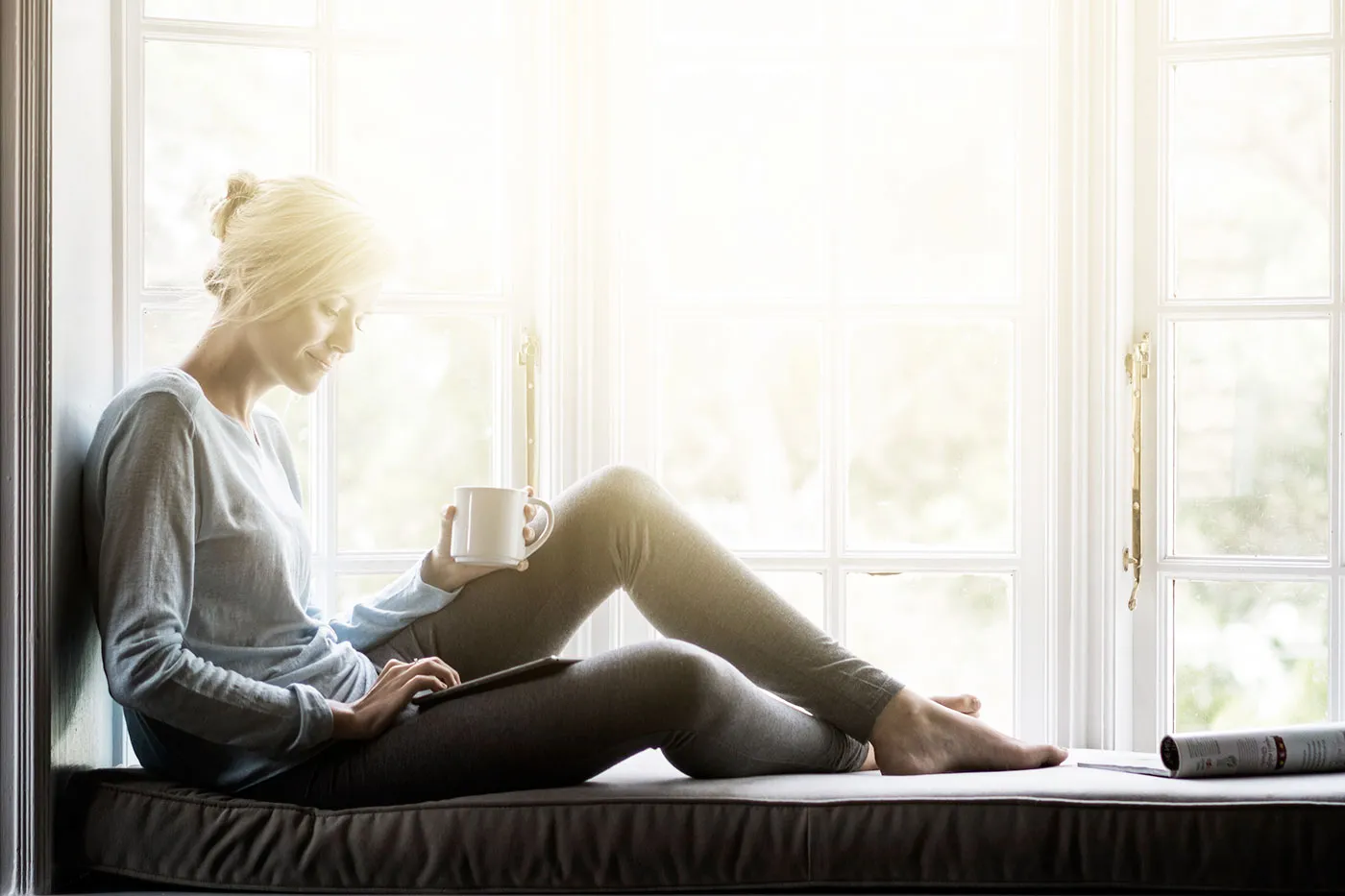 A woman sitting in a sunny windowsill and drinking coffee while looking at her tablet.