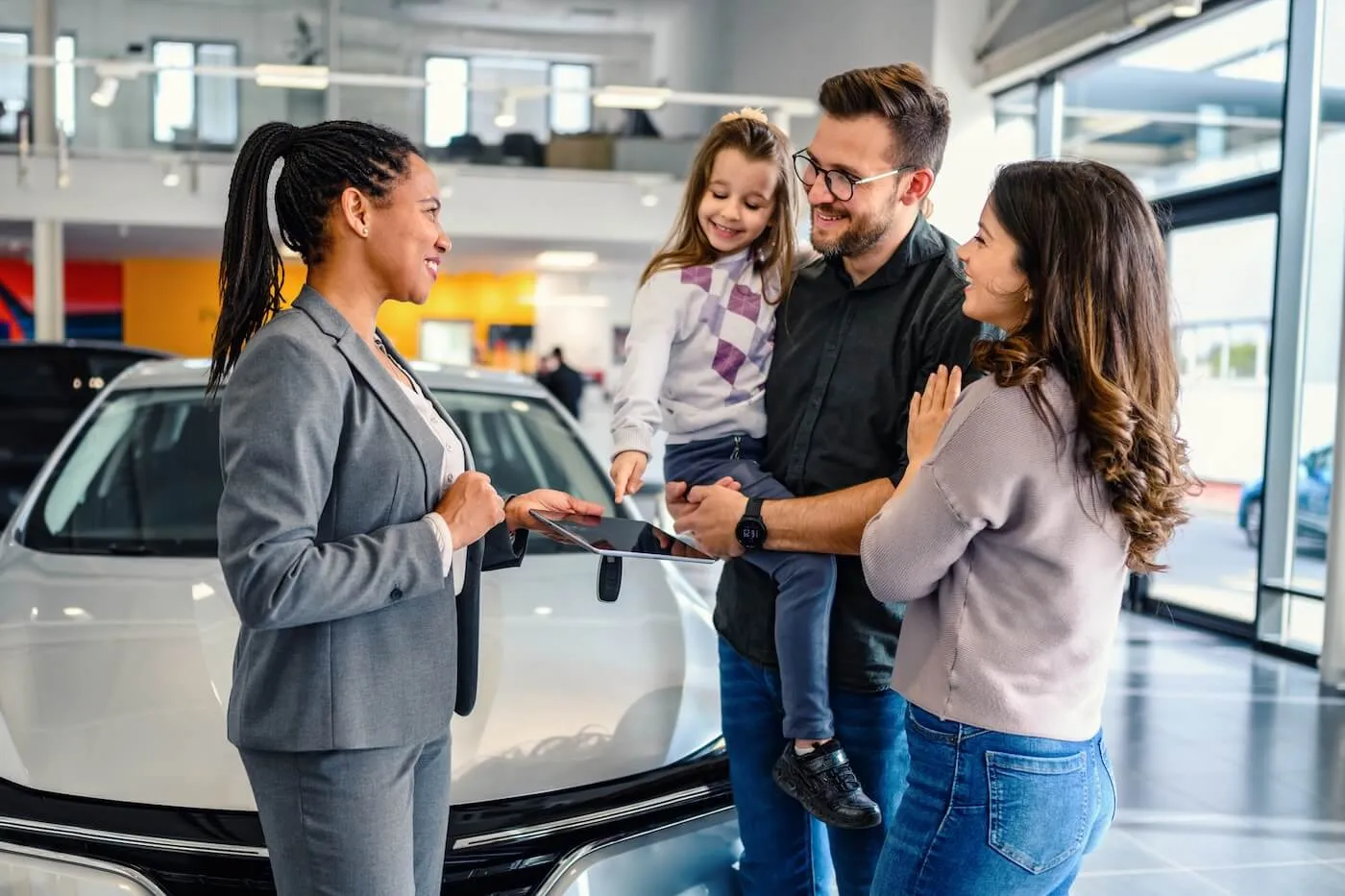 Happy family of three chatting with a female dealership agent who is holding a tablet and car keys. The man is holding a toddler girl. They are standing next to a white vehicle.