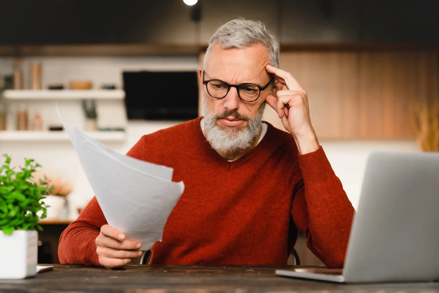 Concerned senior man reviews printouts while using his laptop at home