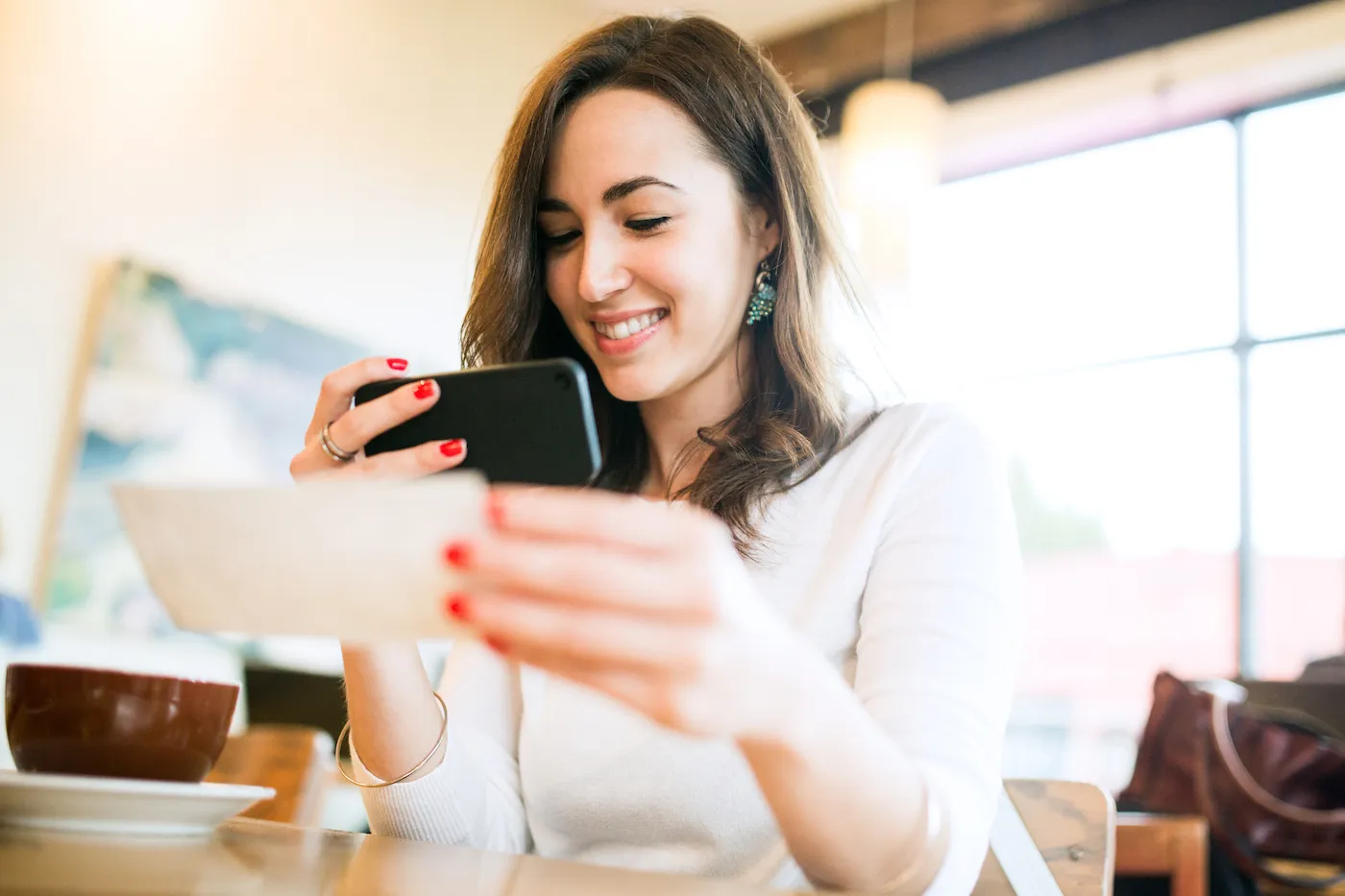 A smiling young woman takes a picture with her smart phone of a check or paycheck for digital electronic depositing, also known as "Remote Deposit Capture". She sits in a coffee shop, enjoying an espresso latte. Bright sunlight shines in the windows behind her.