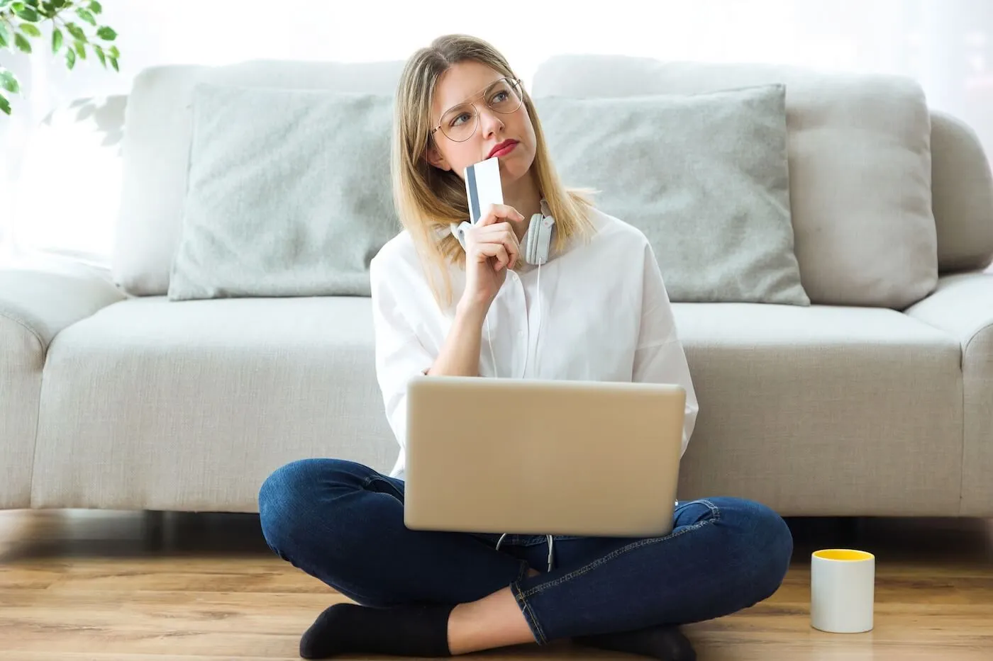 Young woman is sitting on the floor in the living room and thinking while holding a credit card and using a laptop