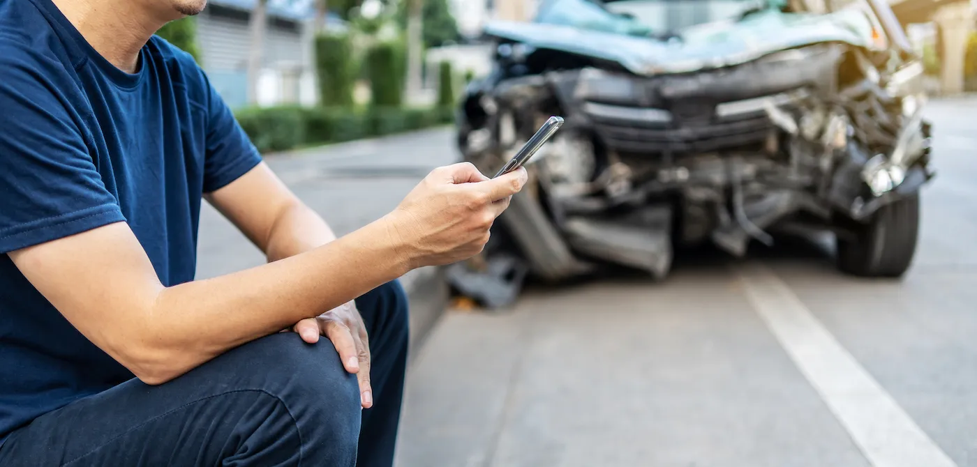 Man calling with phone to his auto insurance agent after a car crash with a background of the crashed car.