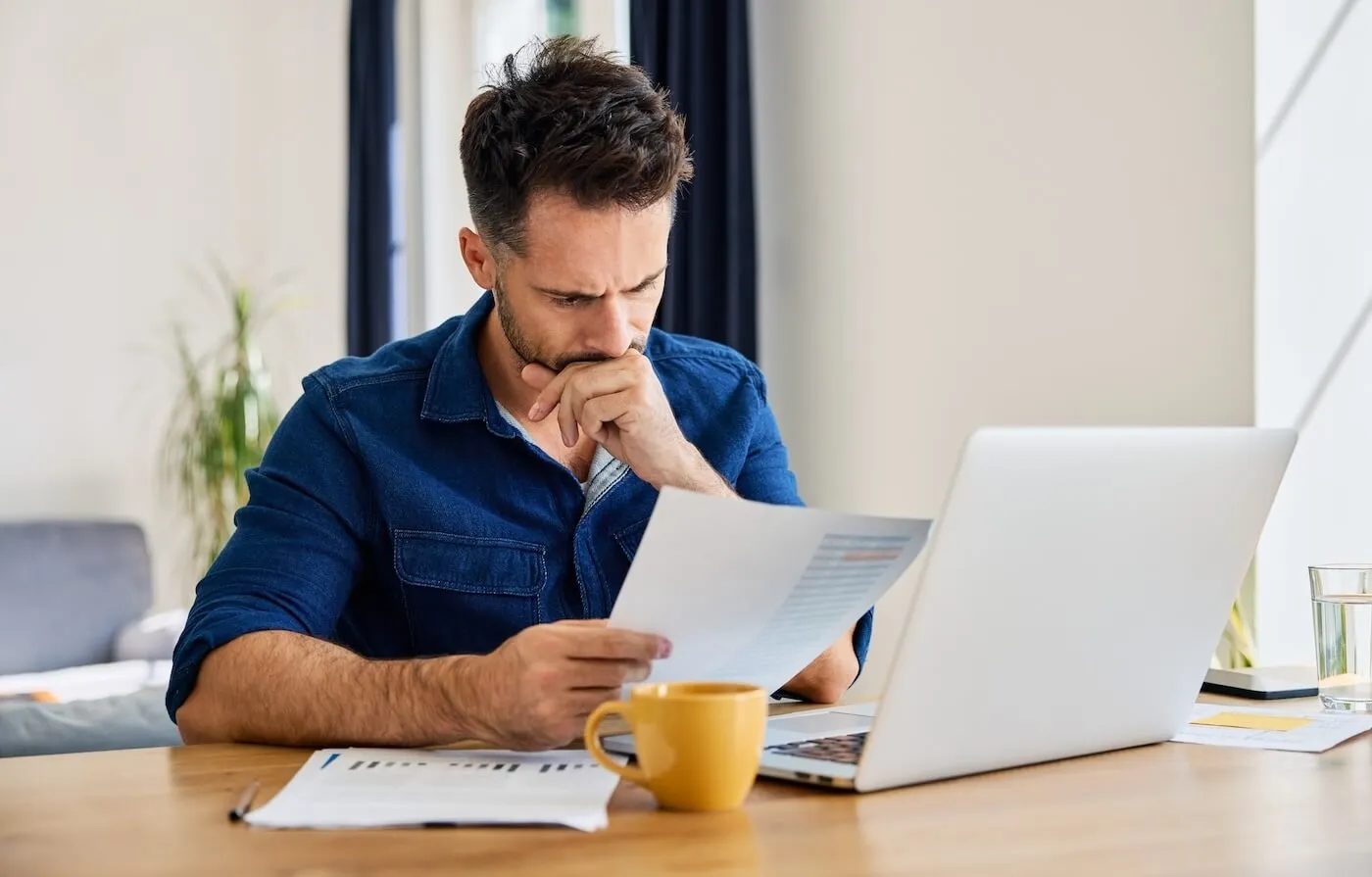 Concerned man reading a printout while using his laptop in a home office, yellow mug standing next to him