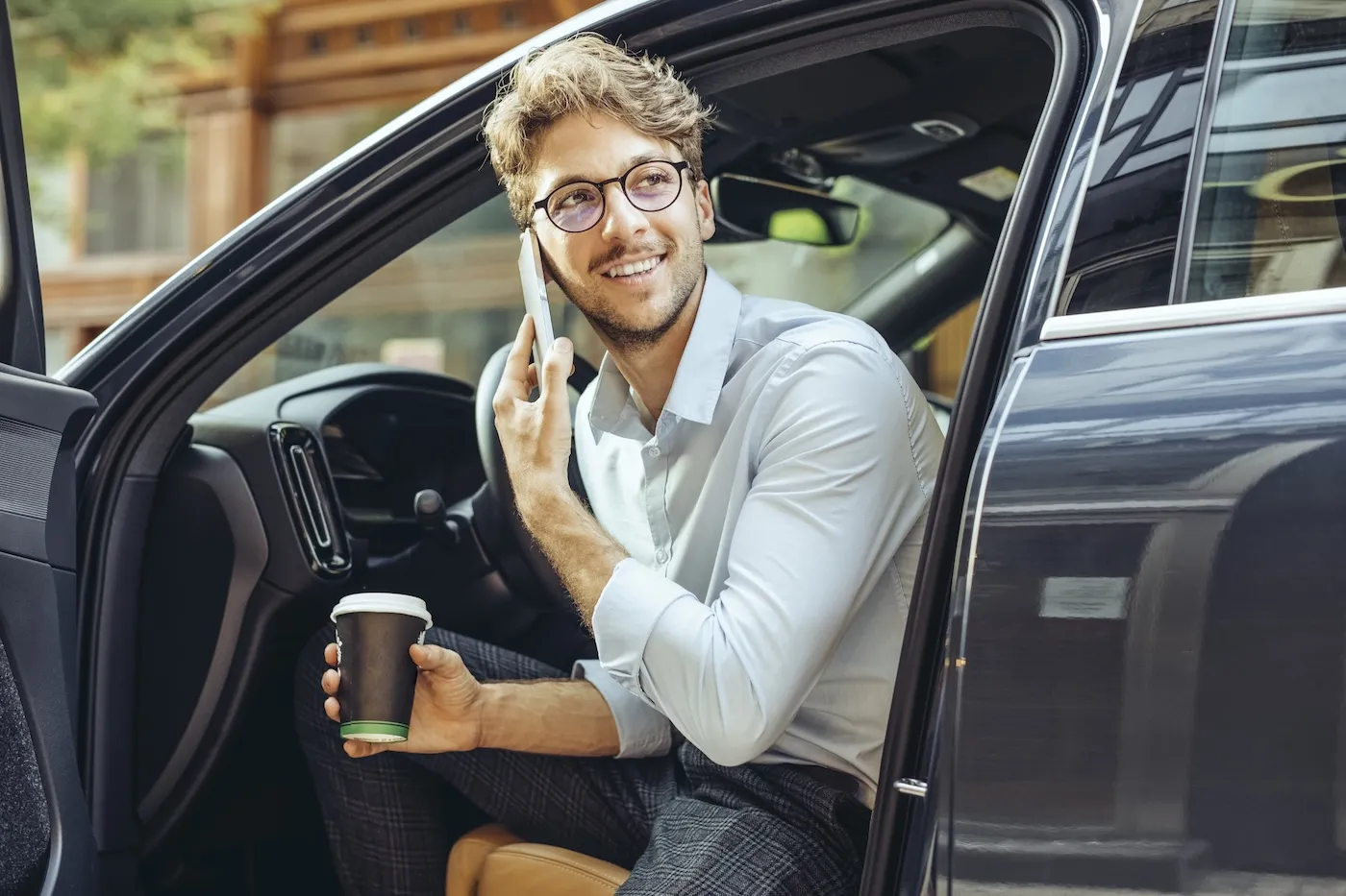 A man talking on the phone and holding his coffee while sitting in the front seat of his parked car with the door open.