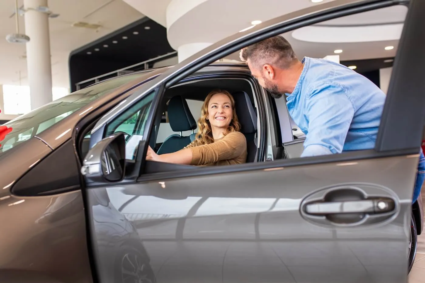 Woman sitting behind the wheel and smiling to a man who opens the car front door in a dealership