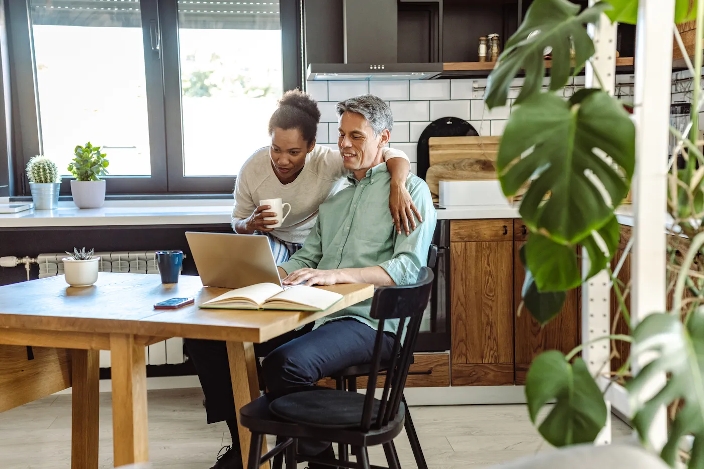 A mature couple is in their kitchen. A mature man is working on the laptop and his wife is next to him.