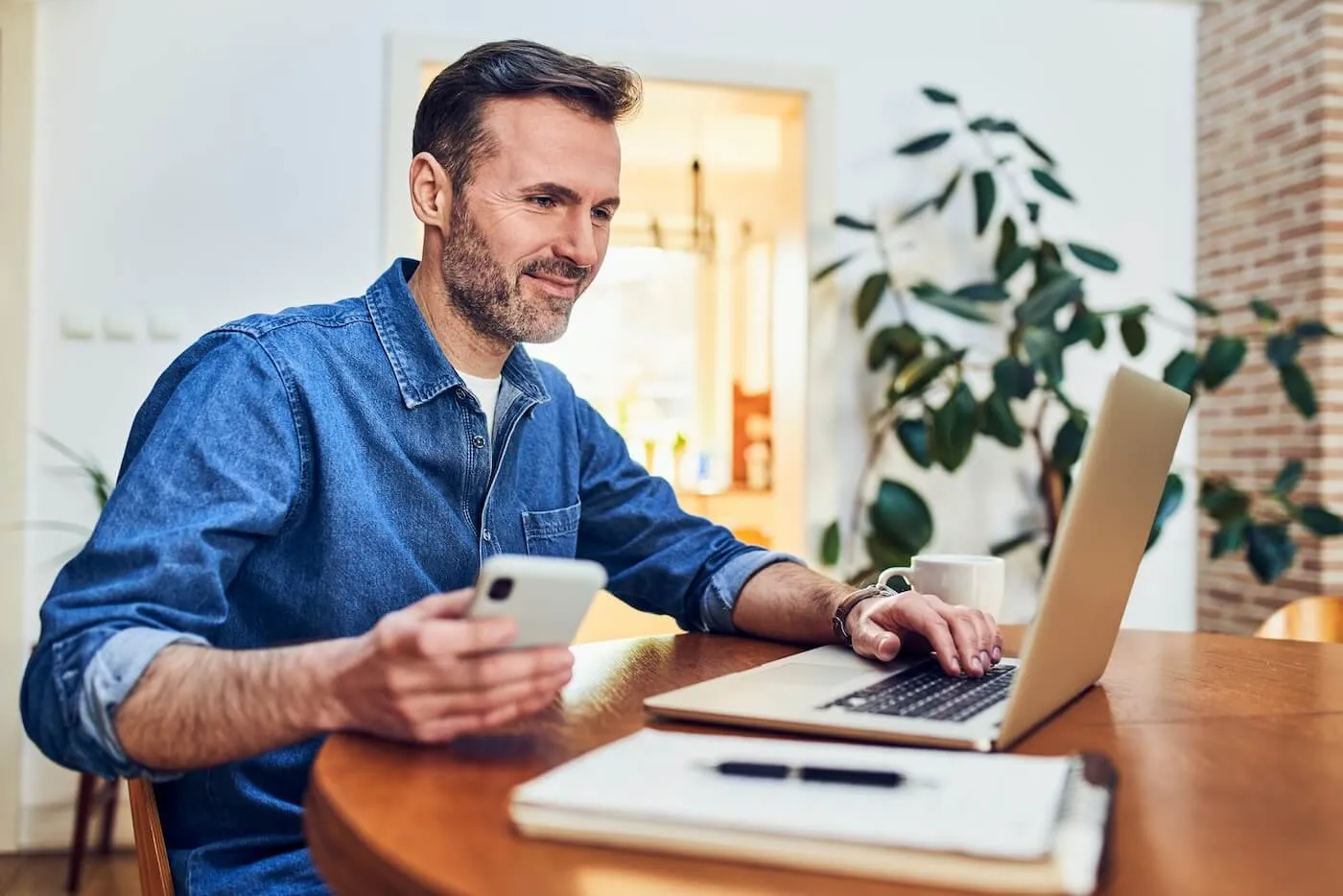 Smiling man using his smartphone and laptop at home