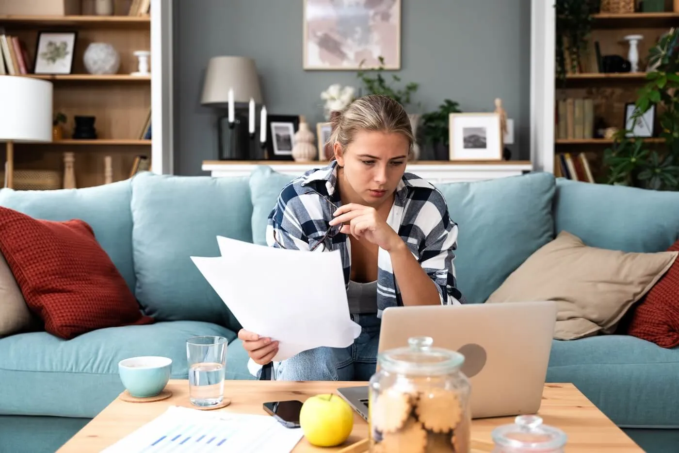 Focused young woman using her laptop while reviewing the papers in her living room