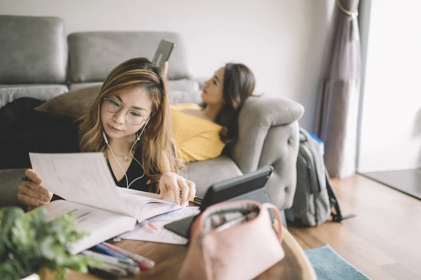 A college student studying in living room, spending time with her roommate who is laying on the couch using her phone.