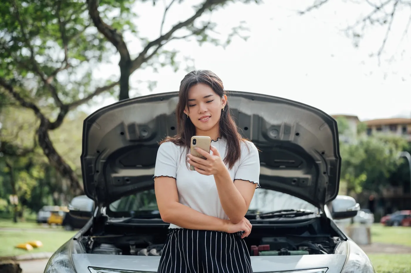 Smiling woman using her phone while standing near her car with open hood on the road side.