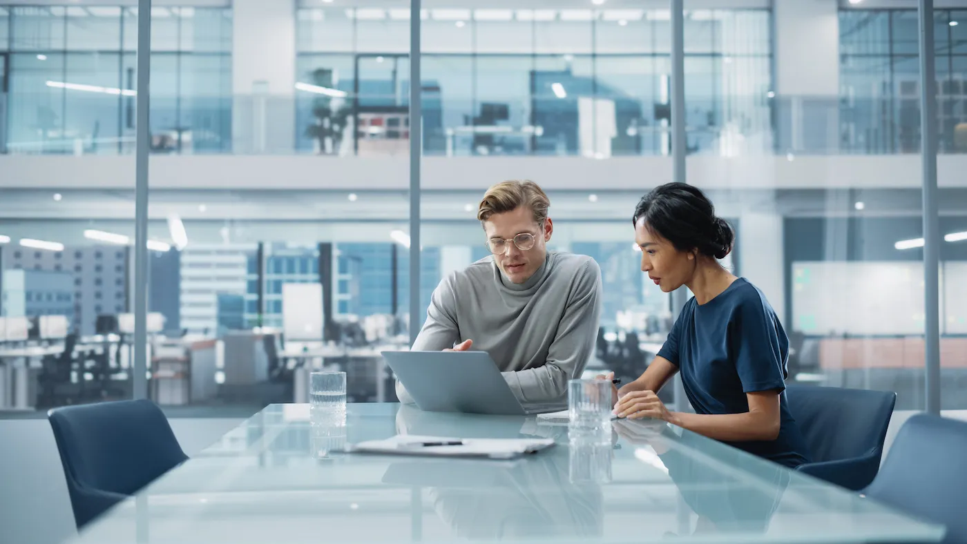 A couple sitting at a large table in an office co-working space, collaborating on a debt repayment plan on a laptop.