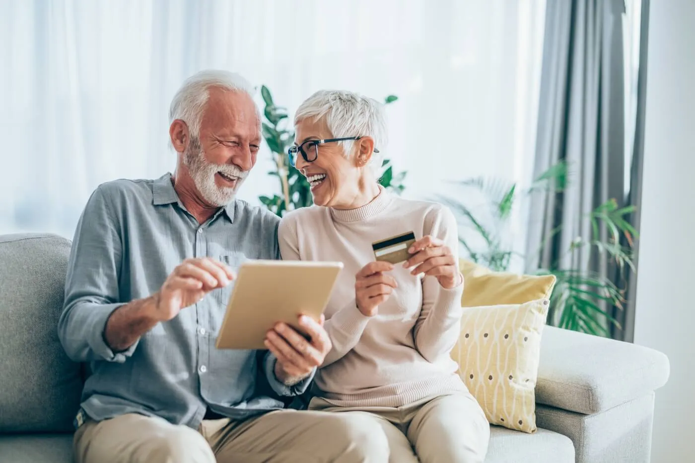 Happy senior couple sitting on the sofa in a sunny room. The man is using a tablet and the woman is holding a credit card.