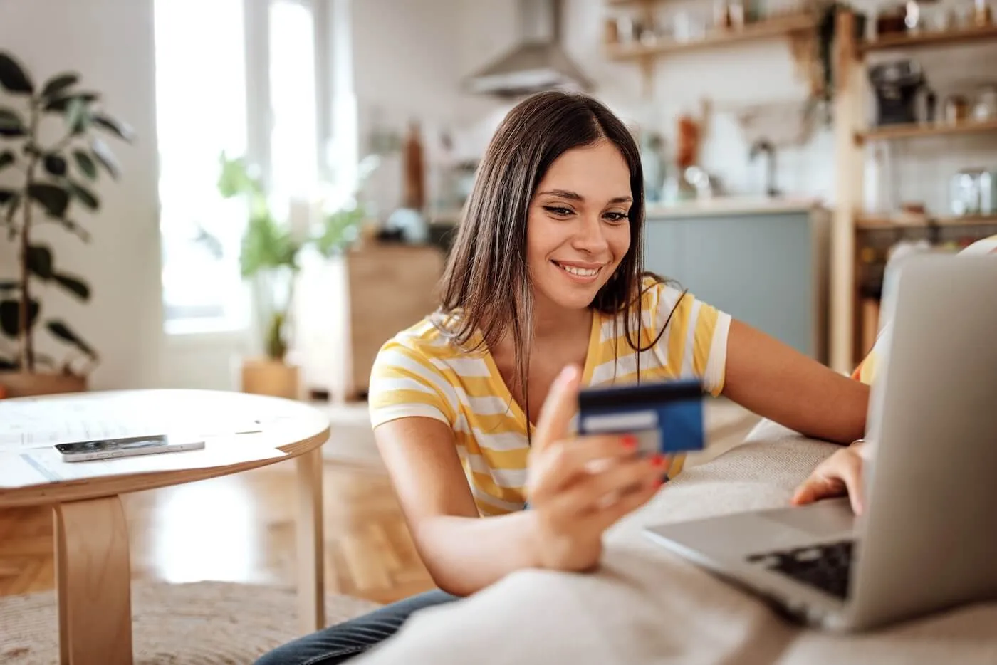 Smiling young woman holding a credit card while using her laptop in the living room