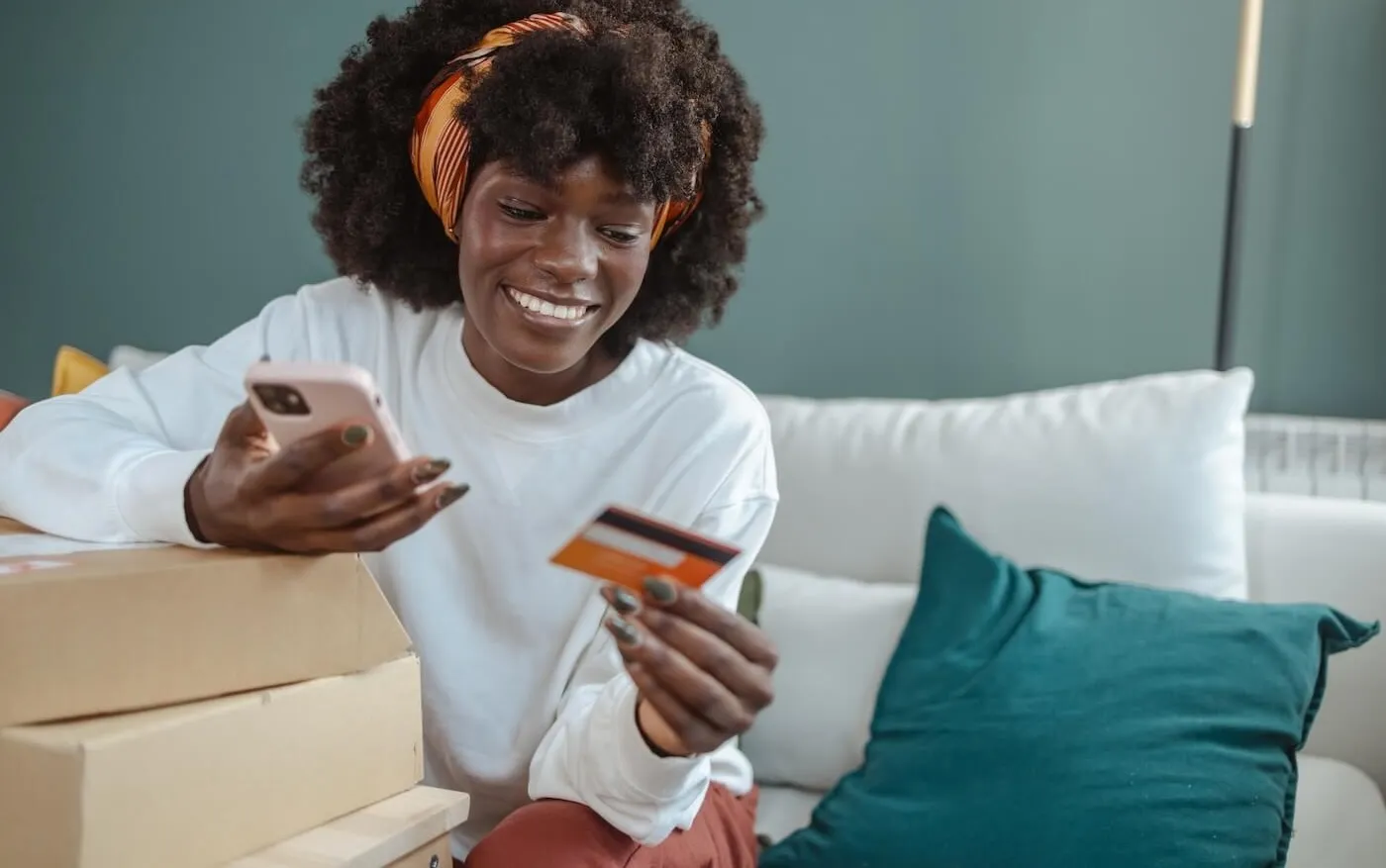 Smiling young woman sitting on the couch with two boxes and holding her credit card and a smartphone