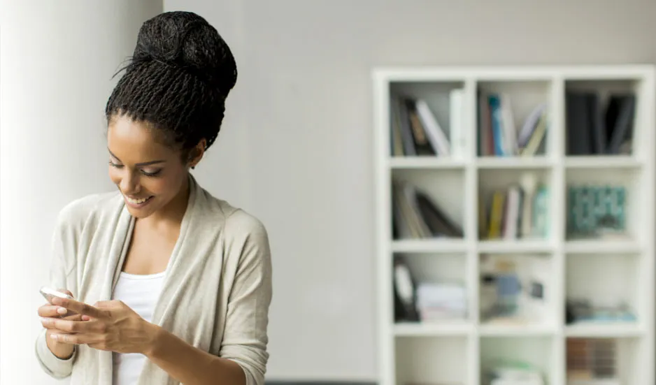 A woman texting and smiling at her phone, standing in front of a white shelving unit.