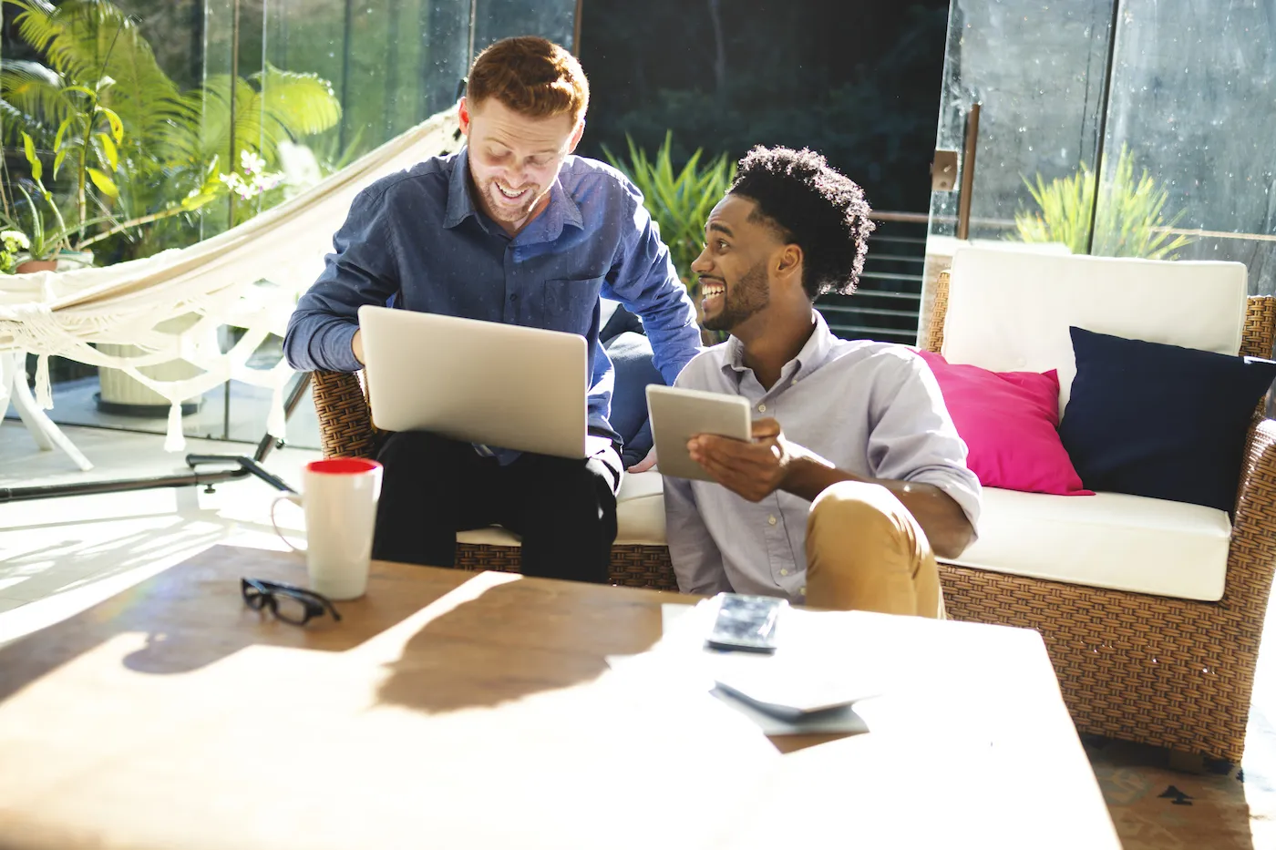 Two young men working at home on their patio, using laptop and tablet while sitting on wicker outdoor furniture.