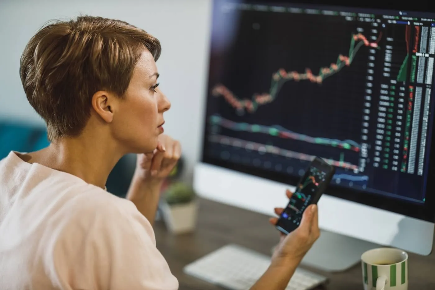 Focused woman sits at a desk holding a smartphone in one hand, with a desktop monitor showing line charts and market data, and a coffee mug nearby