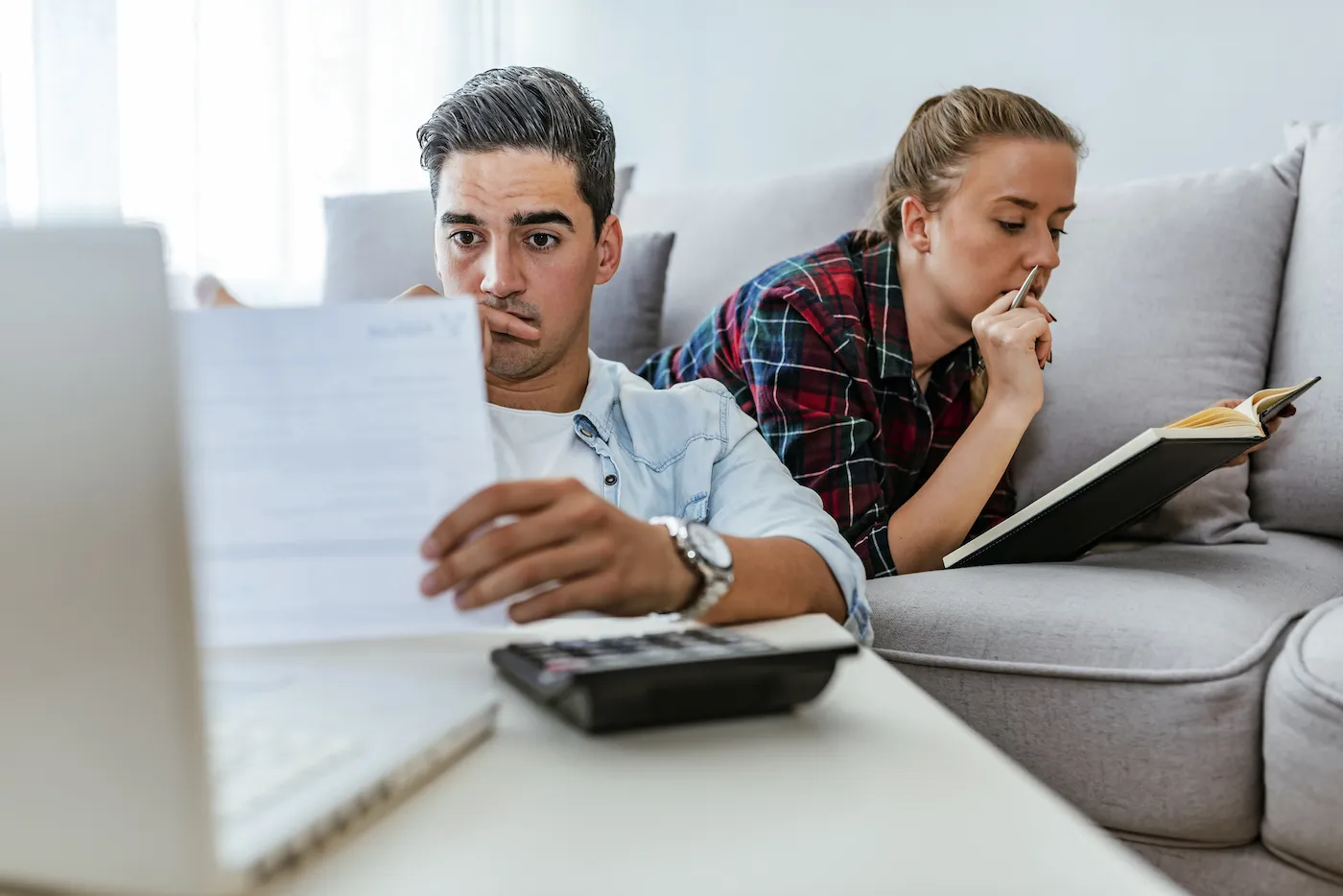 A man looking at his inheritance and wondering whether to share it with his spouse. She is laying on the couch reading a book.