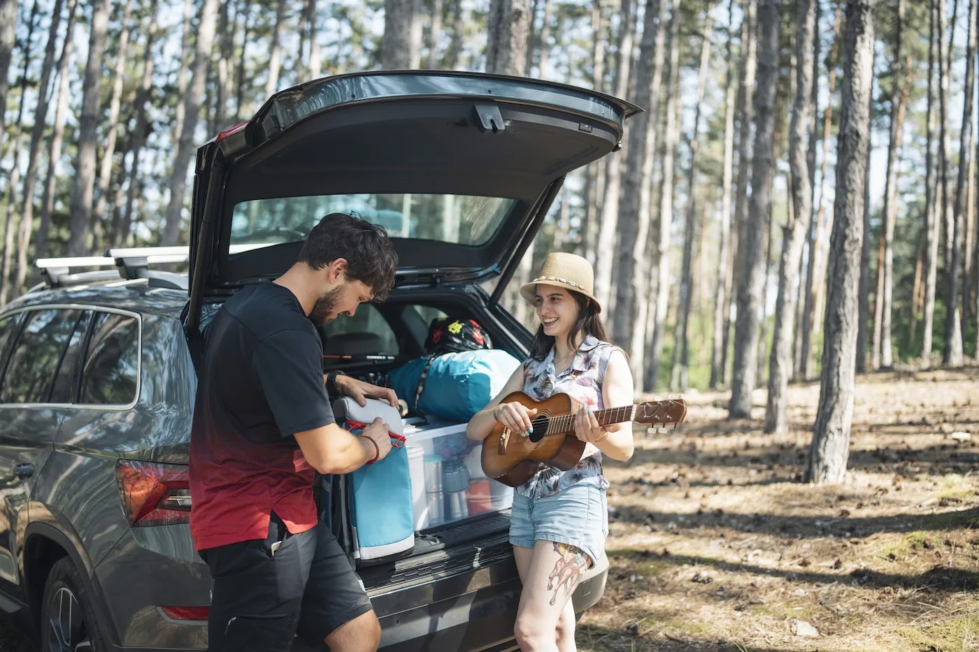 Young couple with their car packed for camping, woman is playing the guitar and man is listening.