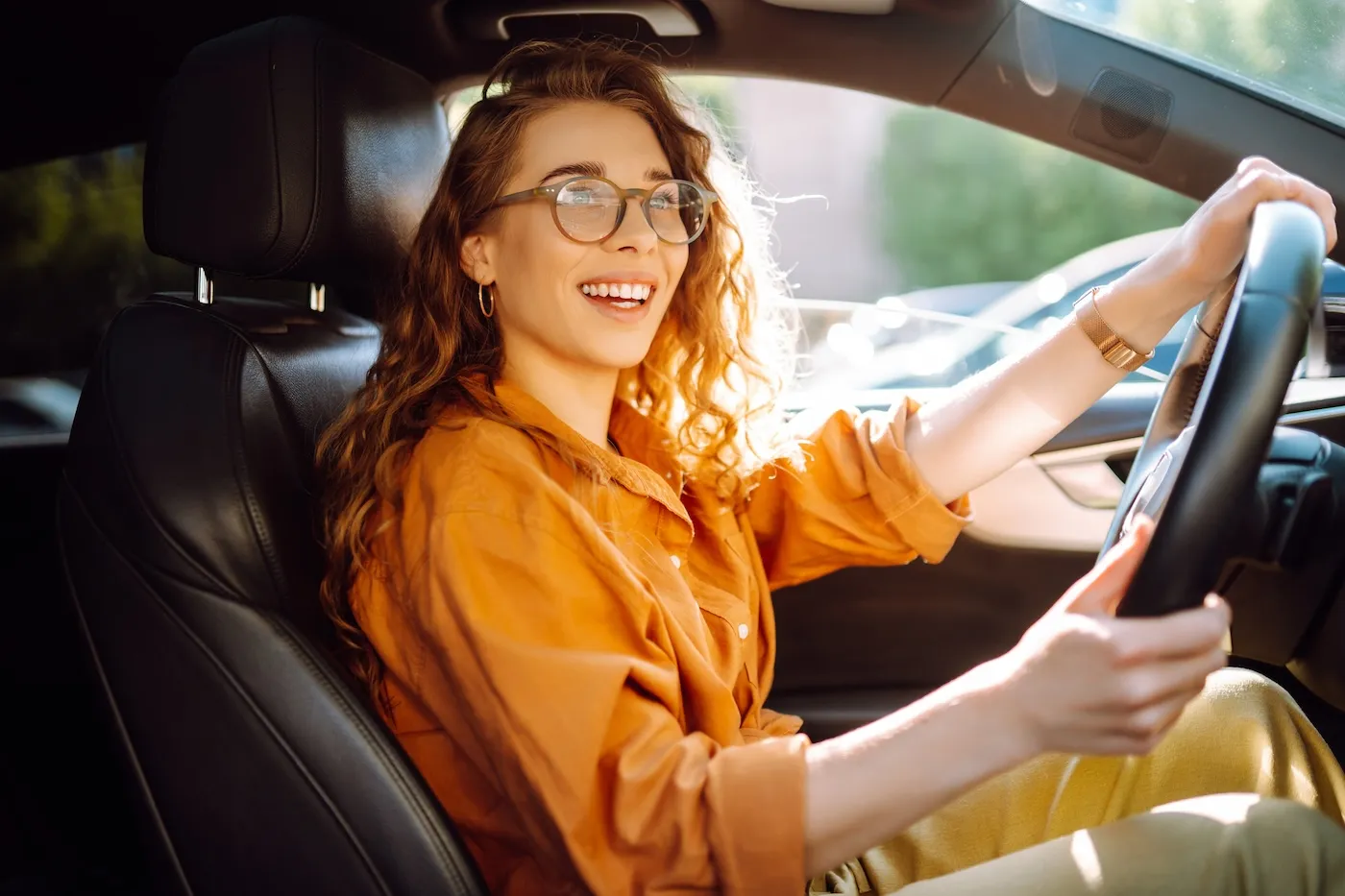 A young woman wearing glasses and an orange shirt driving a car.