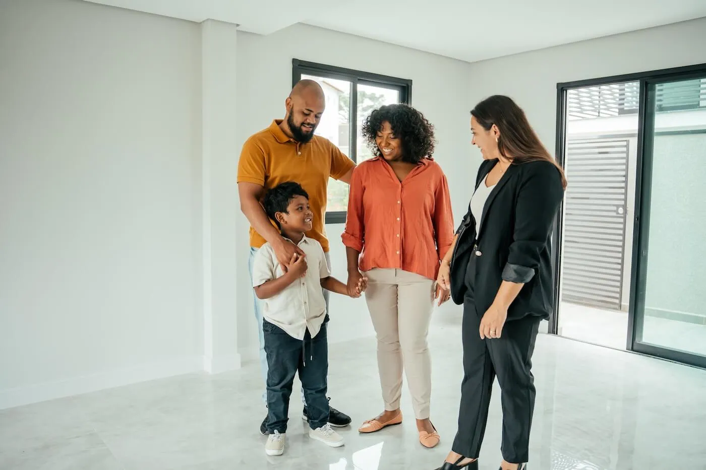 Family of three standing in a bright, empty home with a real estate agent during a property tour. Large windows, sliding glass doors, and open living space are visible as the group walks through the interior