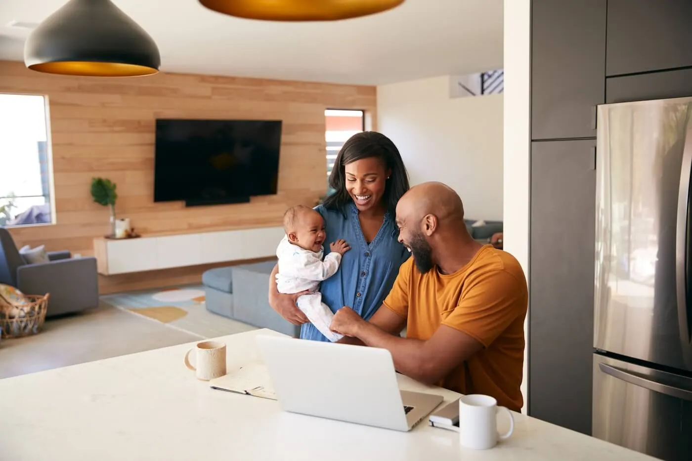 Two adults and an infant gathered around a kitchen island in a modern home, with one adult seated at a laptop and a notebook nearby. Coffee mugs, a smartphone, and a spacious open-concept living area are visible in the background.