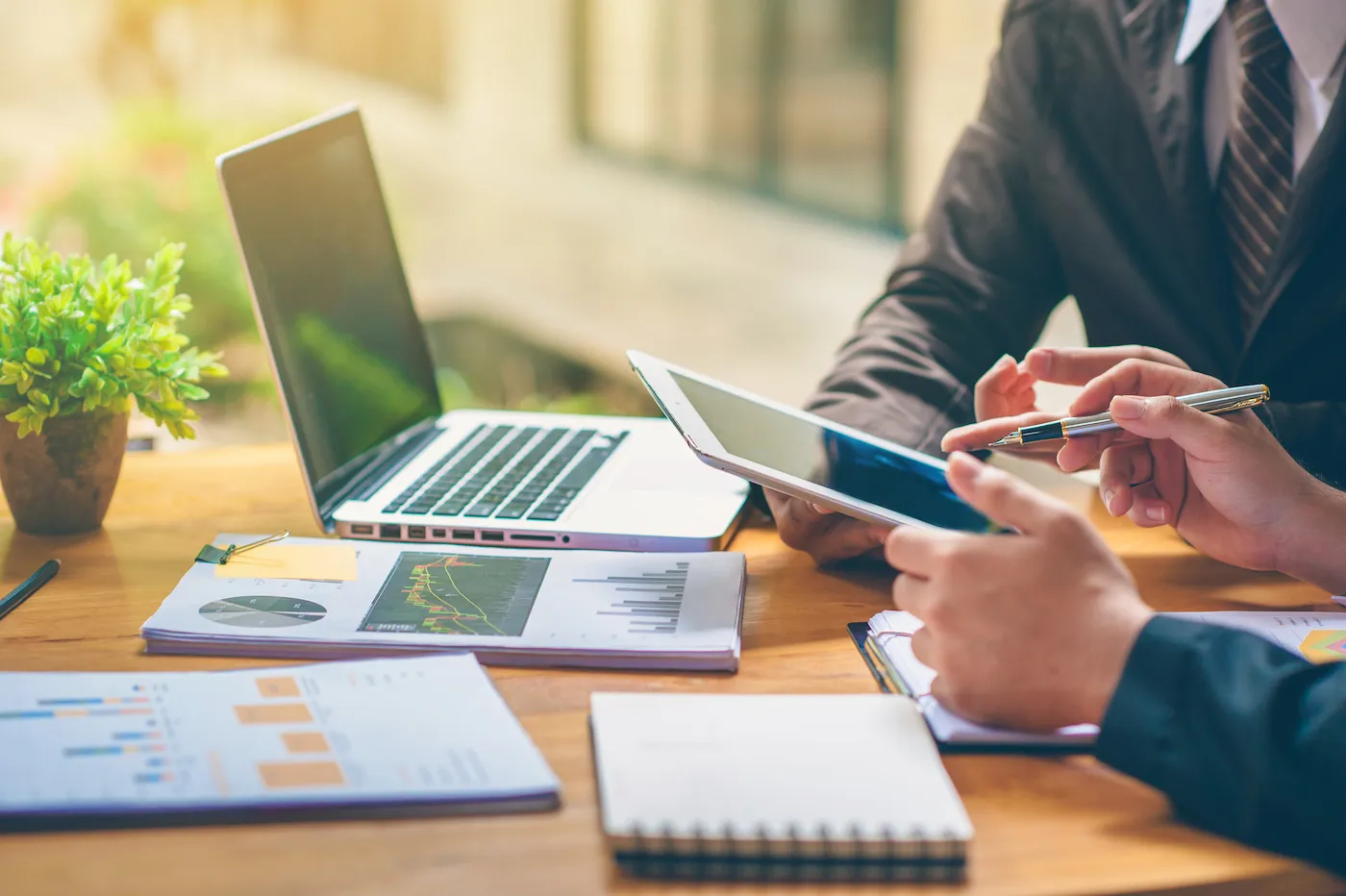 Two professionals sharing finance tips, shot of hands using a tablet and a notepad on a desk with a laptop.