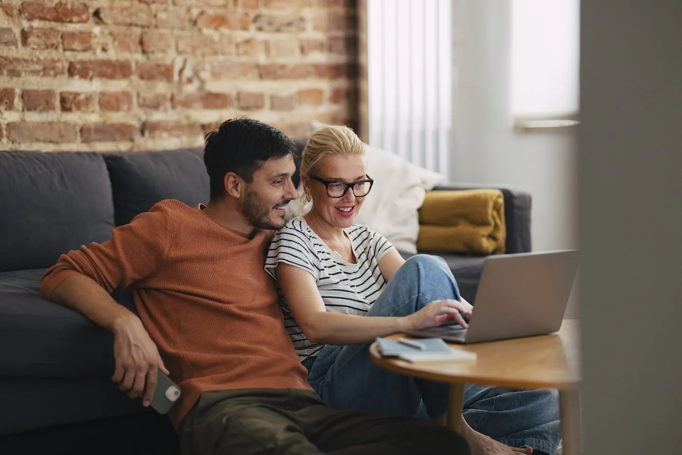 Happy couple sitting on the floor using a laptop computer. They are smiling while looking down at the screen.