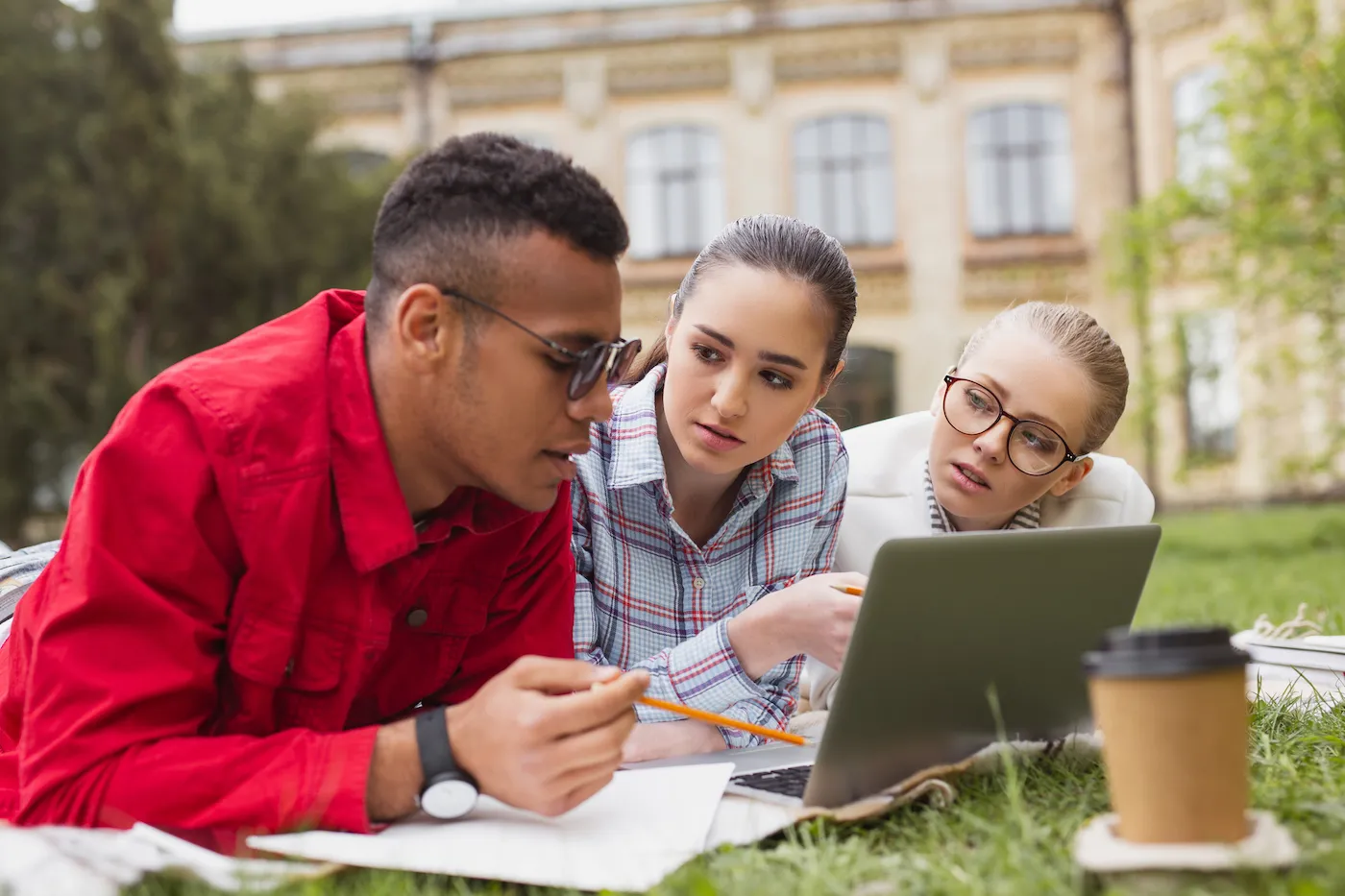 Three students researching credit card interest rates, laying on stomachs on the campus lawn with a laptop.