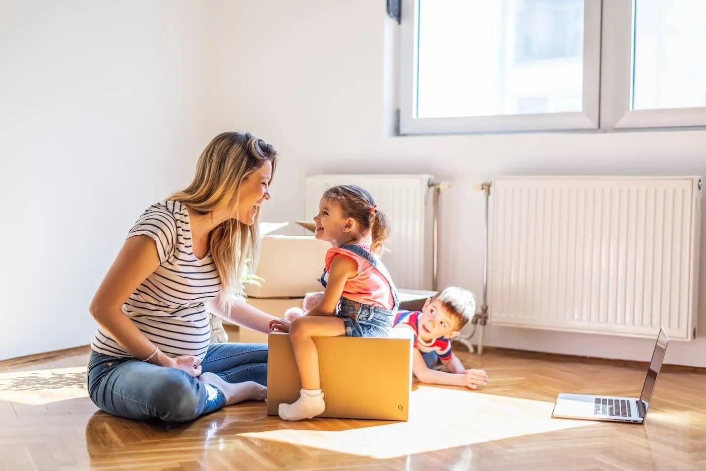 Pregnant woman is sitting on the floor of the empty and sunny living room and smiling to a toddler girl who is sitting on a moving box. A young boy is looking at them while lying on the floor and using a laptop