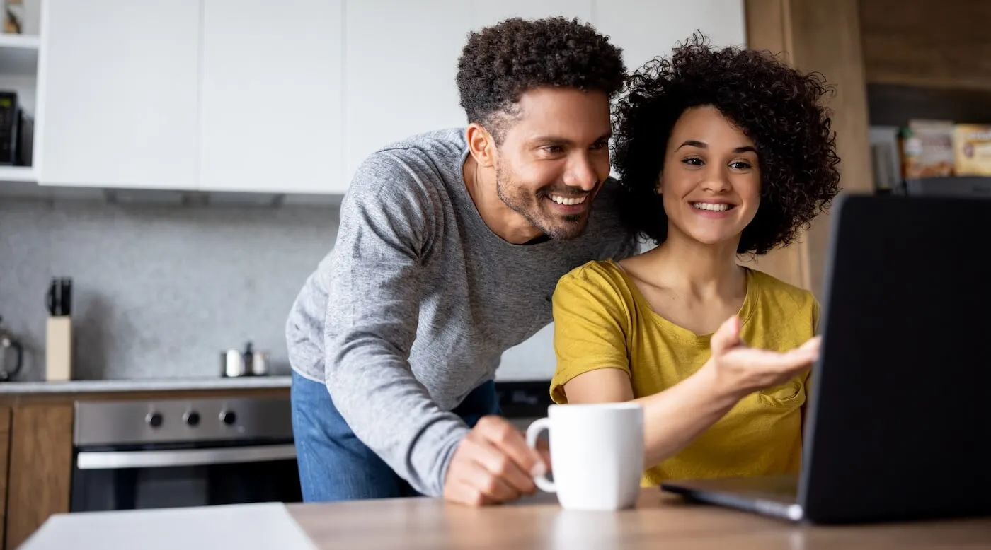 A smiling woman is showing something on a laptop to her partner while having a coffee