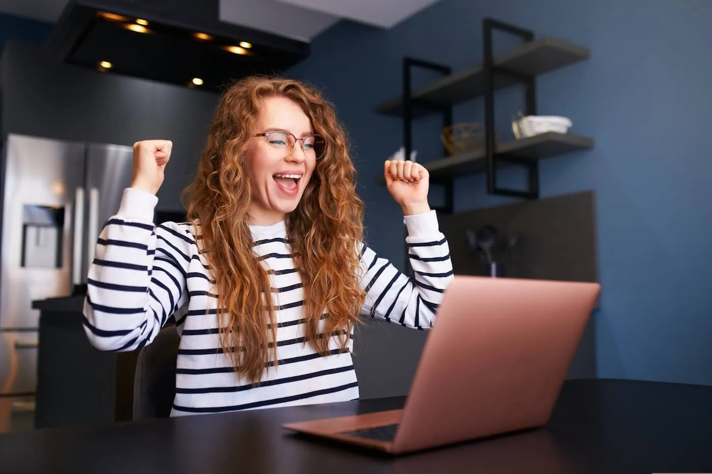 Excited woman sitting at a desk in a modern kitchen, raising both fists while looking at a laptop with a wide open-mouth expression; wearing glasses and a striped long-sleeve shirt, with shelves and kitchen appliances visible in the background