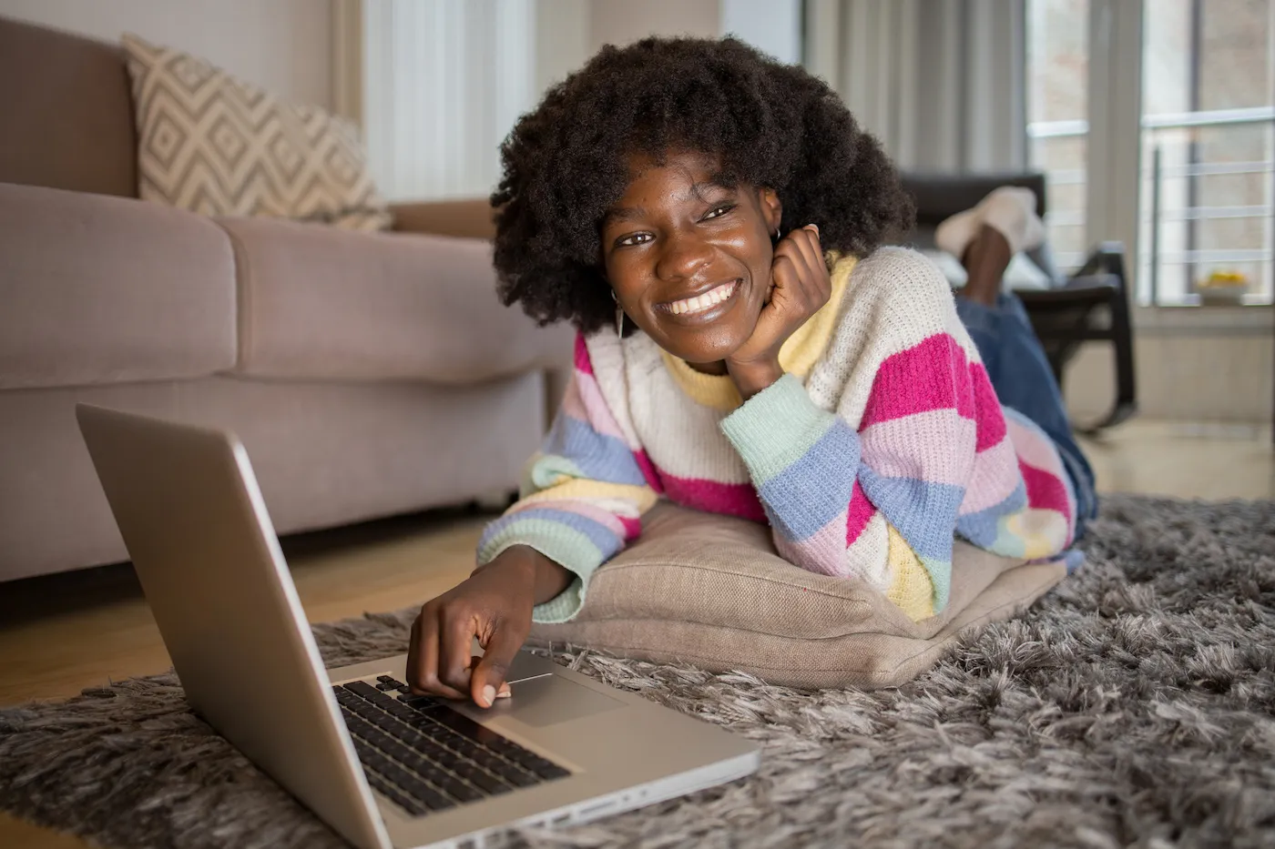 Happy college student wearing colorful striped sweater is laying on her stomach on the ground, using her laptop to research financial accounts