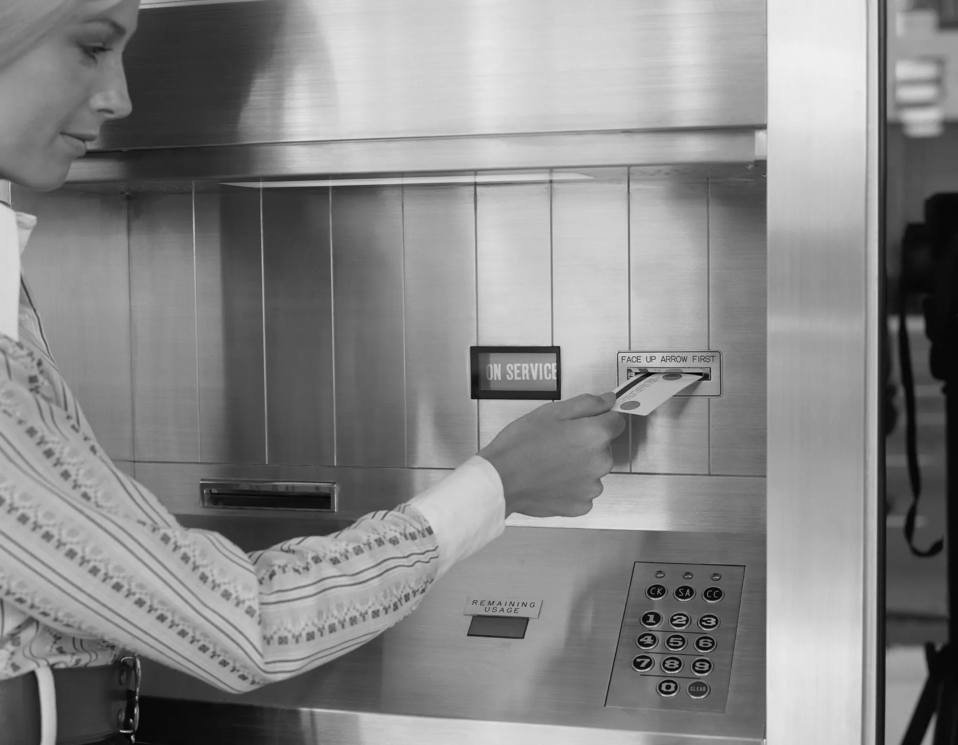 A vintage black-and-white photo of a woman entering a credit card into cashpoint.