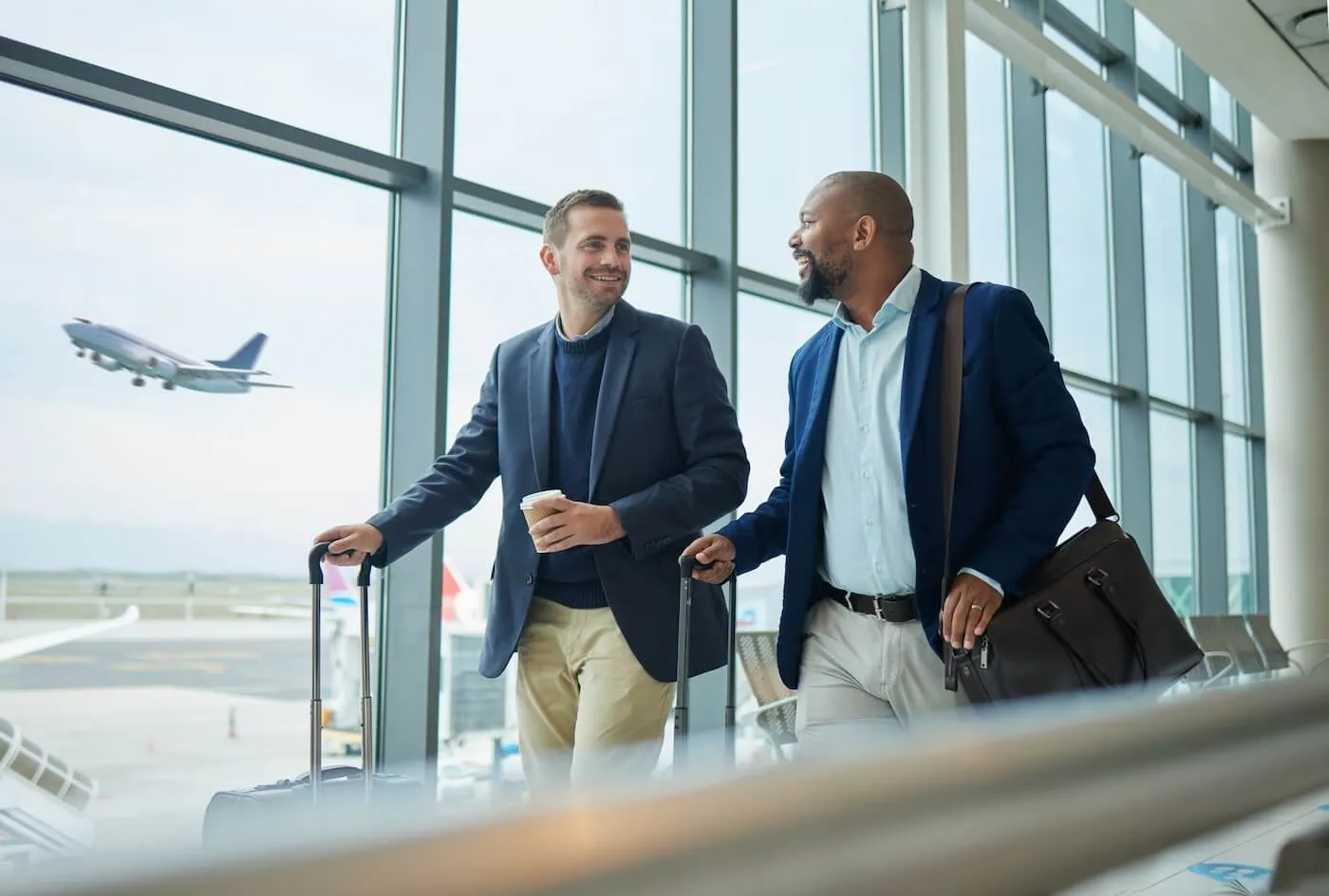 Two men wearing suites are walking to the airport gate with their luggage while the plain is taking off on the background