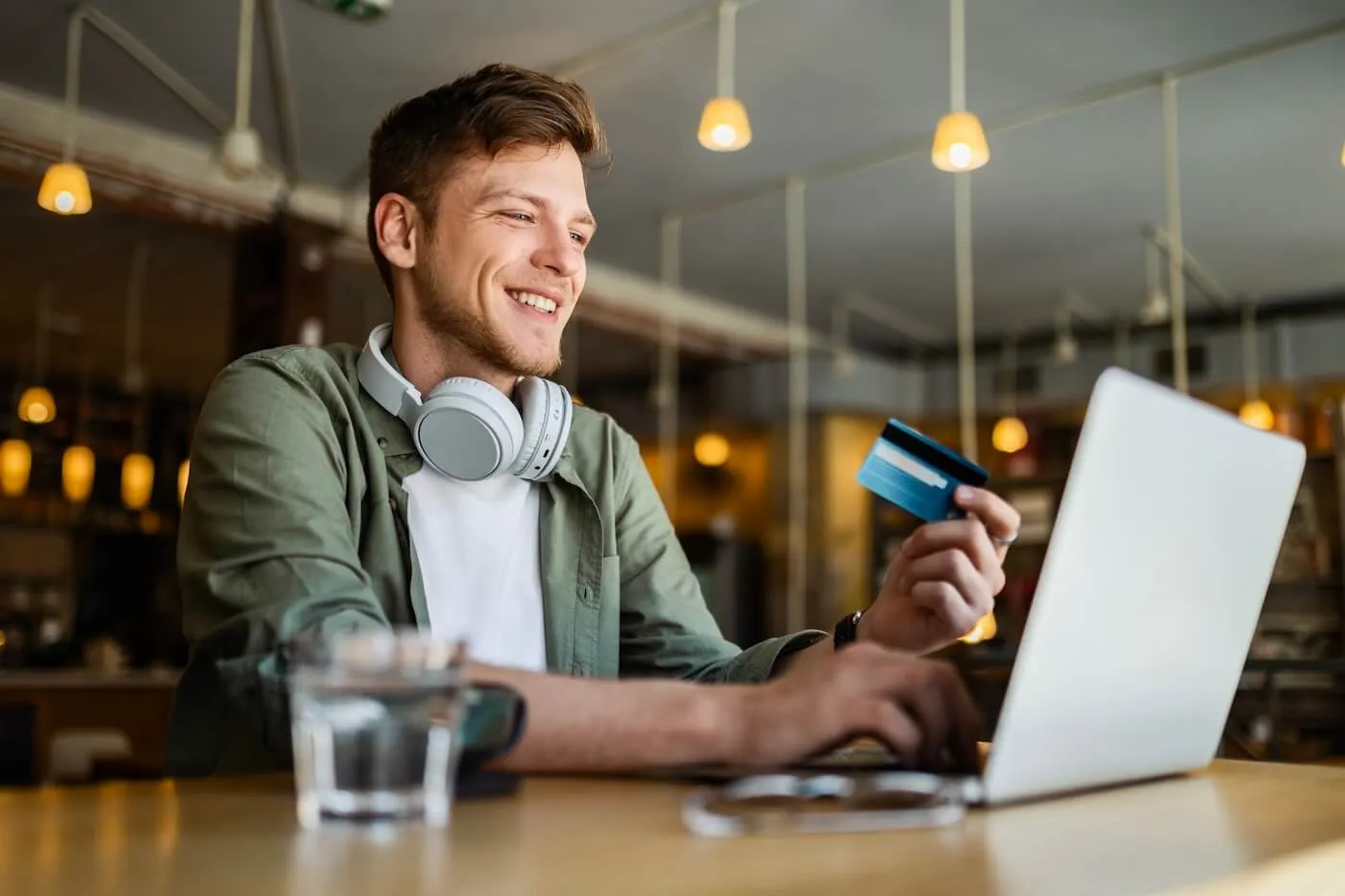 Smiling young man with headphones is holding a credit card while using his laptop in the cafe